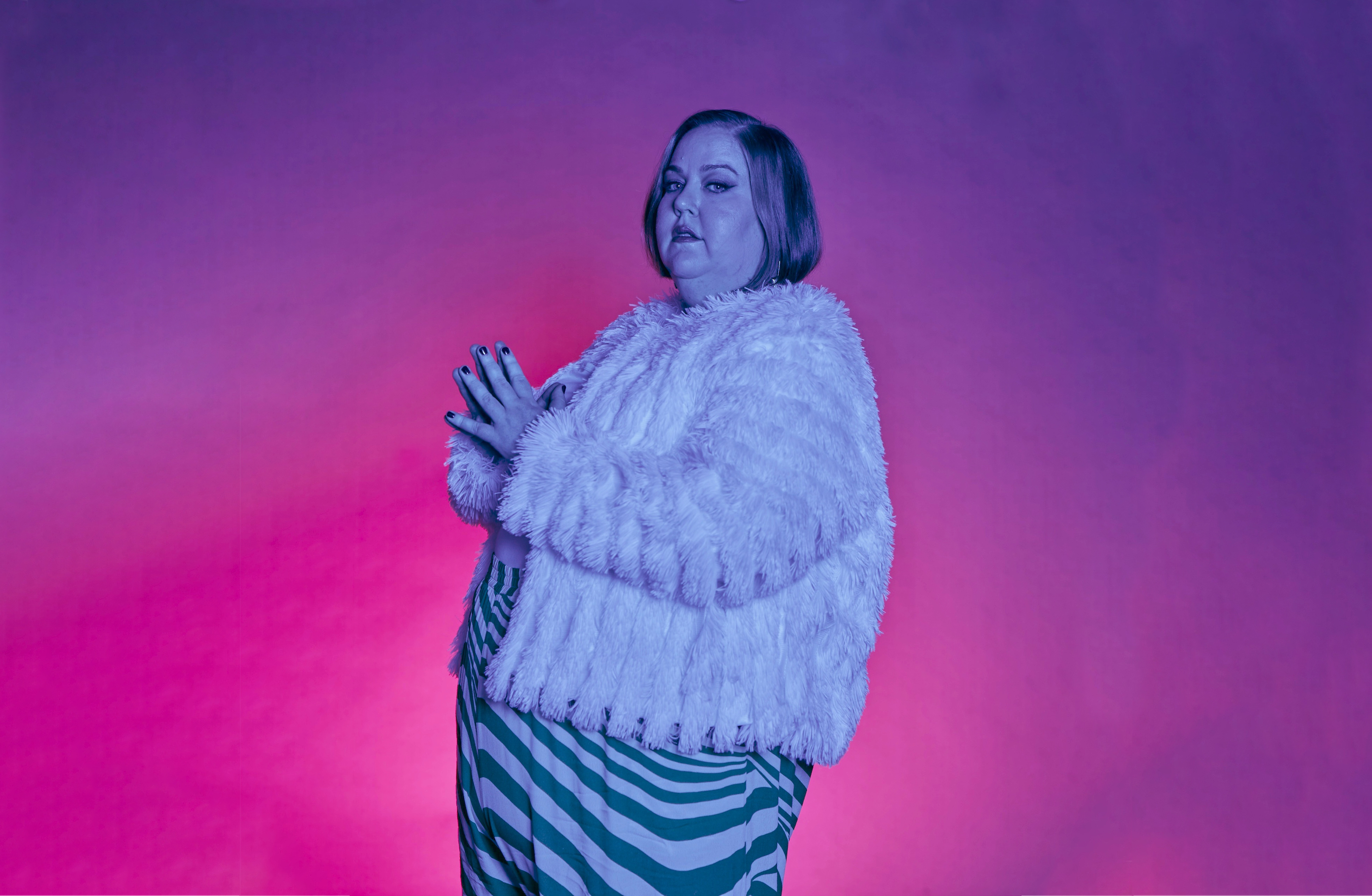 Still from Your Fat Friend. A fat woman (Aubrey Gordon) with blonde hair is viewed from the side, wearing a striped dress and a fluffy jacket whilst bathed in bi-sexual pink and blue light and set against a pink and purple background.