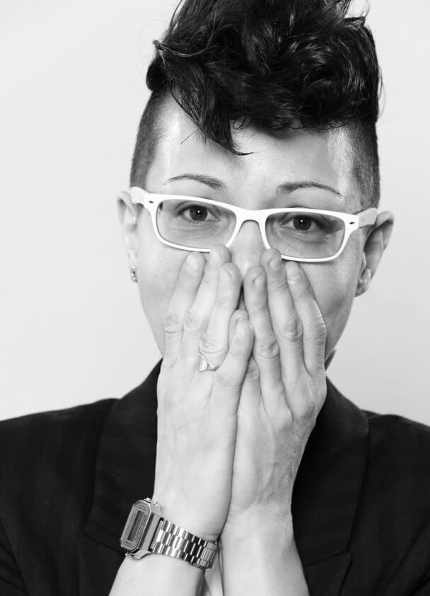 Ilinca Calugareanu looking directly at the camera, smiling. She is wearing glasses and her hands cover her mouth. Black and white portrait