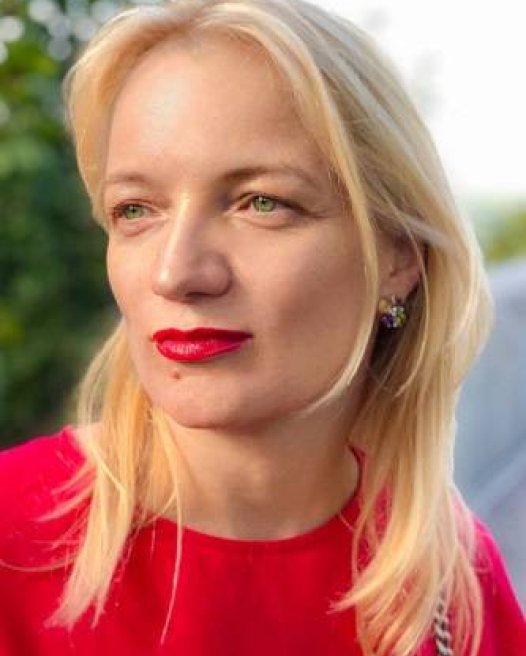 Anna Kapustina, a blonde woman with a bold red lip, looks away from the camera.