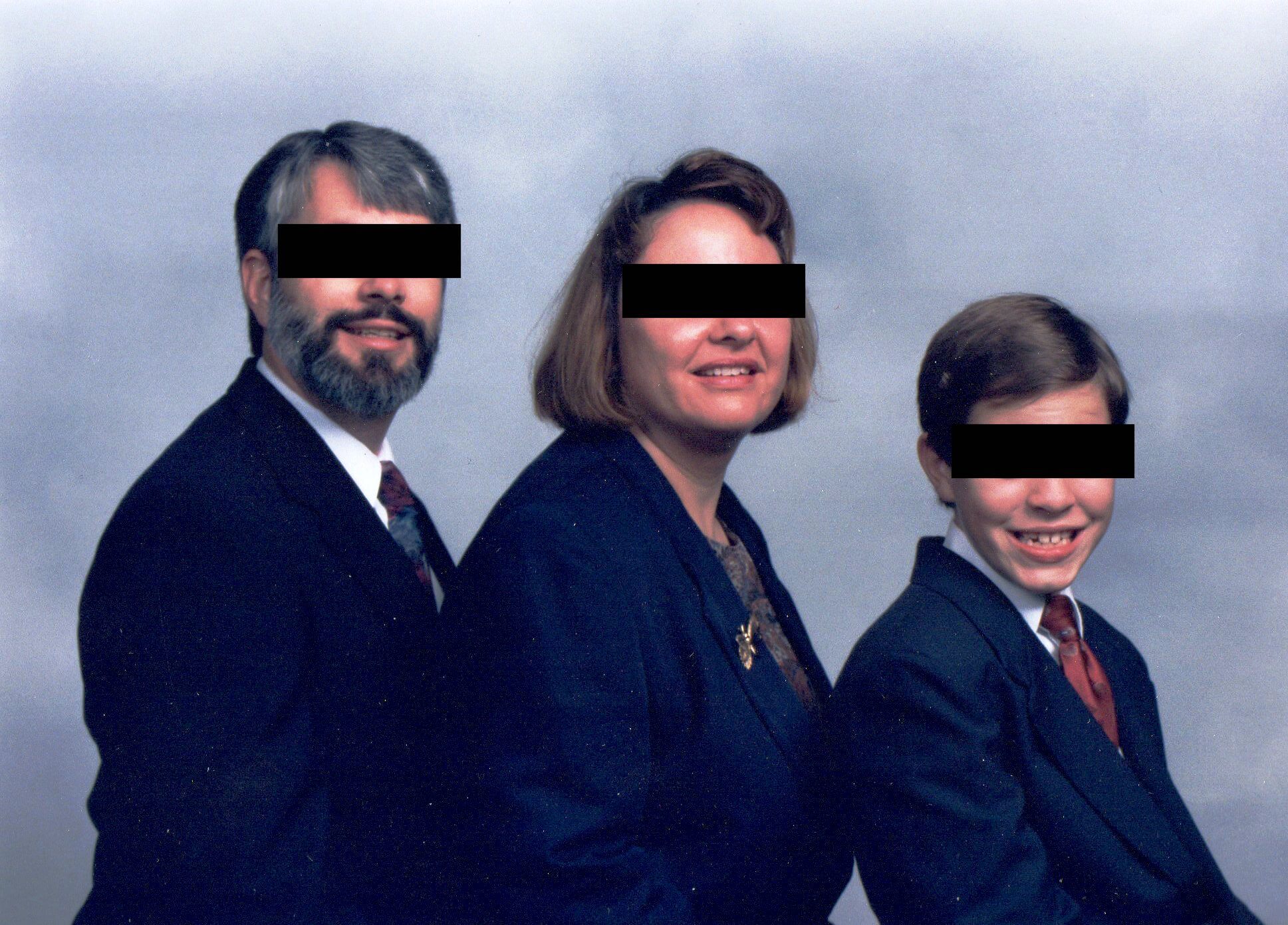 A family portrait of a man, woman and child all of whom wear suits or blazers. They are facing the camera but their identity is partially obscured by a black box covering each of their eyes. 