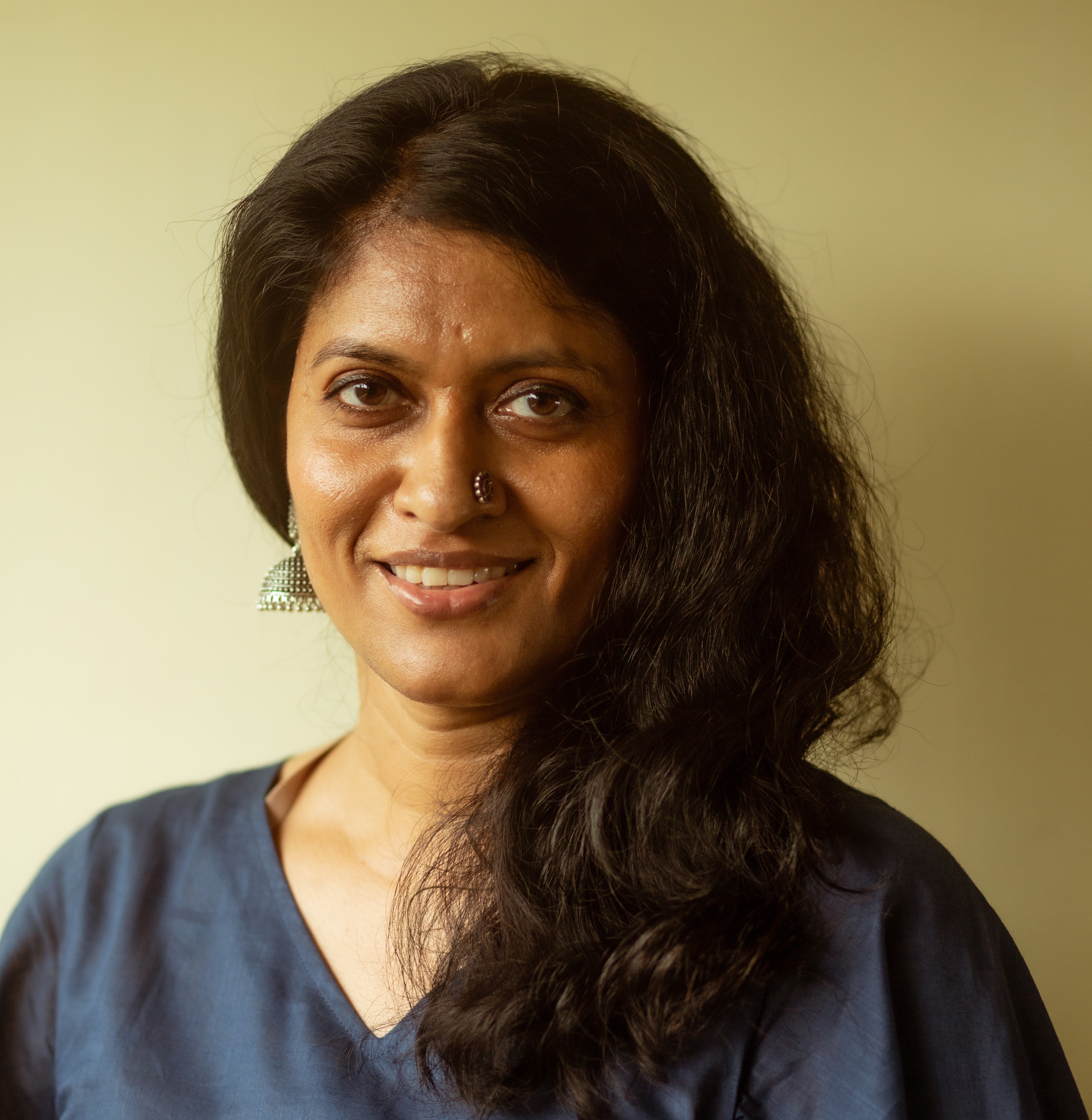 A warm color portrait of Chithra Jeyaram, a South Asian filmmaker, smiling directly at the camera. She's wearing a navy blue top, a stone-studded nose ring, and large semi-dome-shaped silver earrings. Her shoulder-length dark brown hair is gathered on her left shoulder.