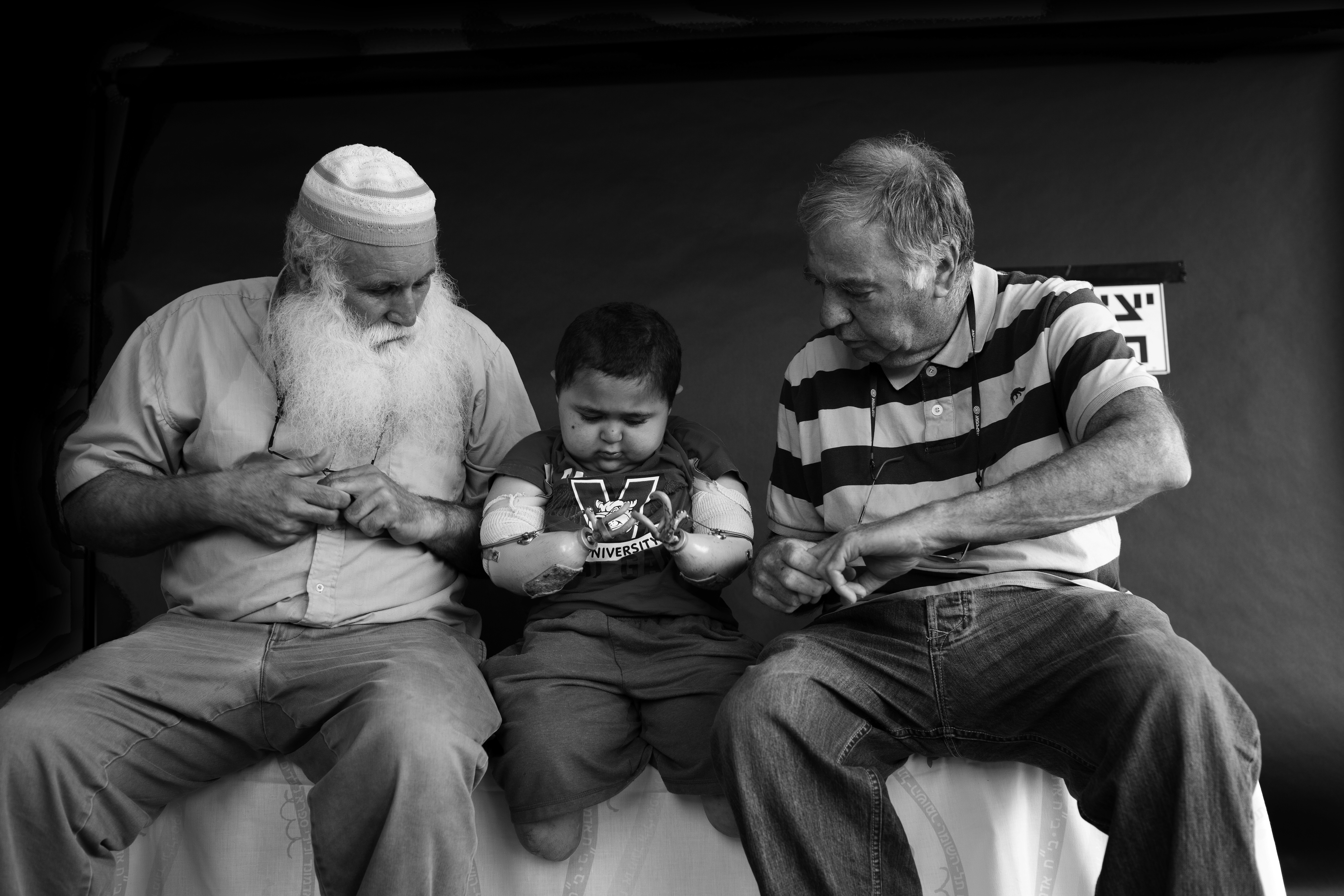 Two men sit on either side of a young child with prosthetic arms and bandages on, and they gesture with their hands.