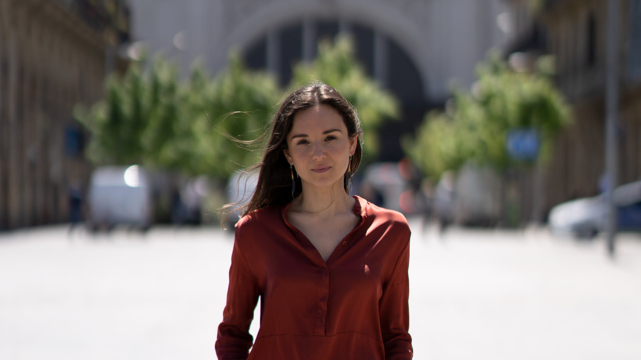 Headshot of Ainara Vera from the waist up wearing a long sleeve dark blouse standing in front of a blurred building and trees.