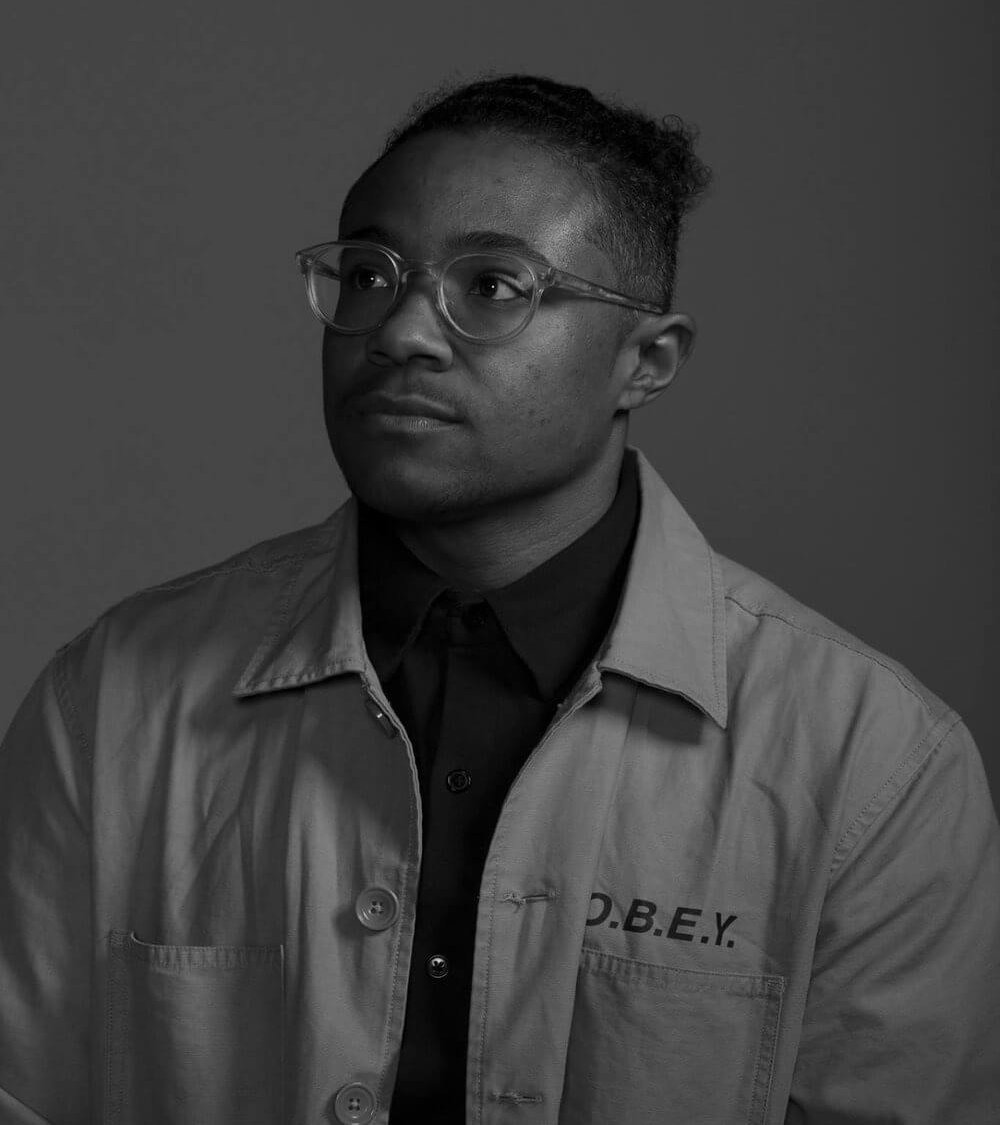 A black man wearing his hair in a bun and a tan chore jacket looks off frame.