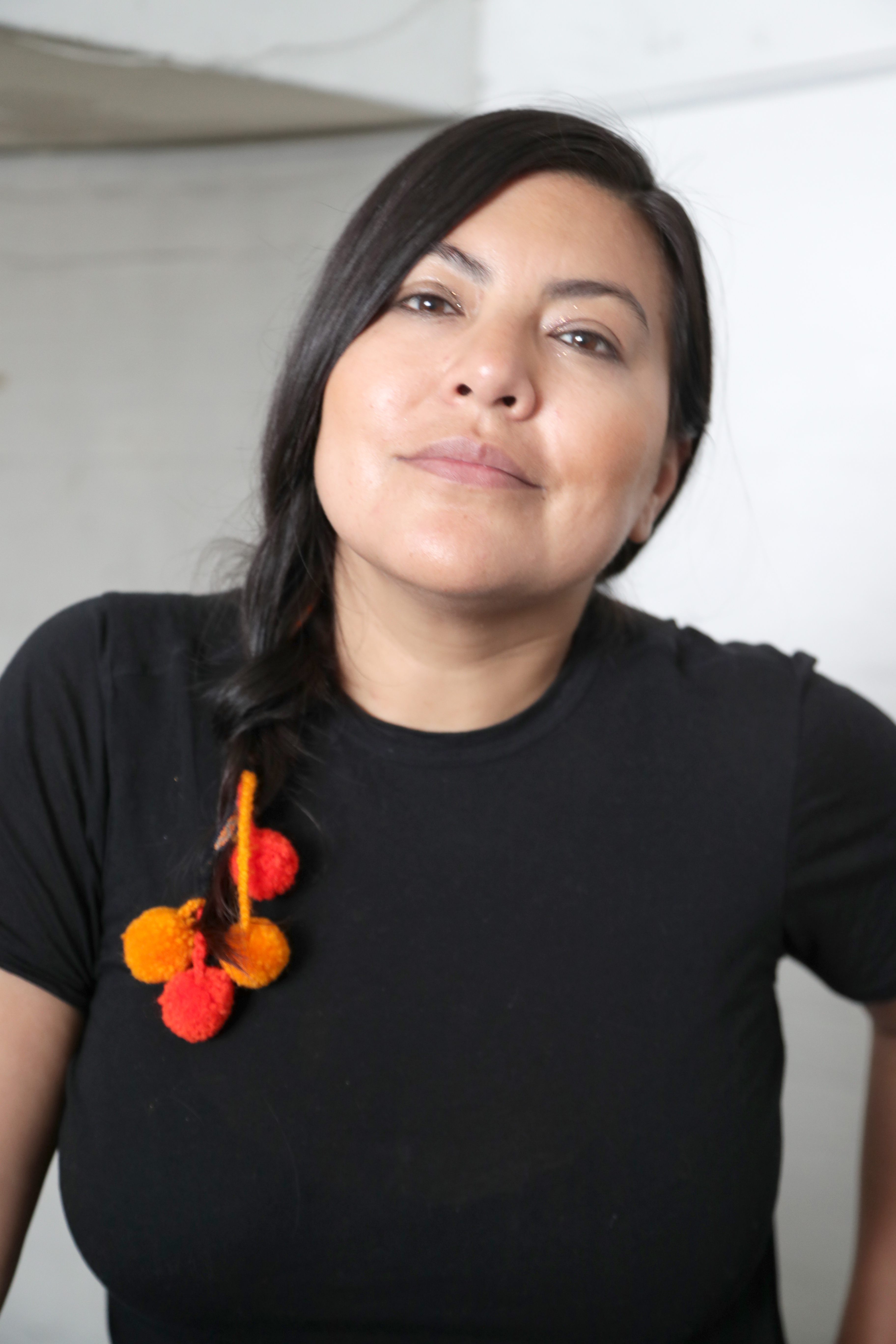 Violeta Ayala is shown in a close-up portrait. She has straight, dark hair styled in a braid, adorned with colorful red and orange tullmas. Her expression is calm and confident, highlighting her strong features, including high cheekbones and a warm, light brown complexion. She is wearing a black shirt, and the plain background keeps the focus on her face, reflecting her Quechua heritage.