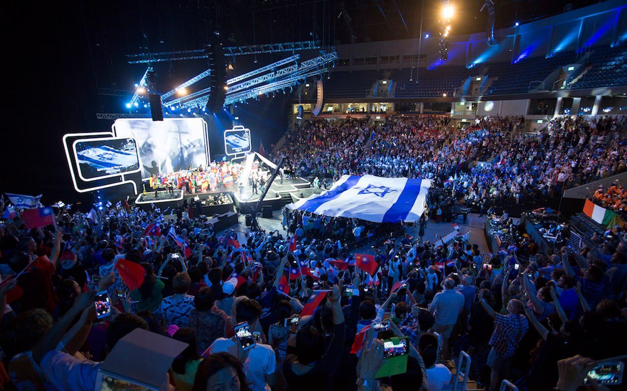 Still from Praying For Armageddon. Evangelical conference hall with thousands of people in front of main stage. Projections of cross on stage. Huge Israeli flag stretched out in the middle of the crowd.