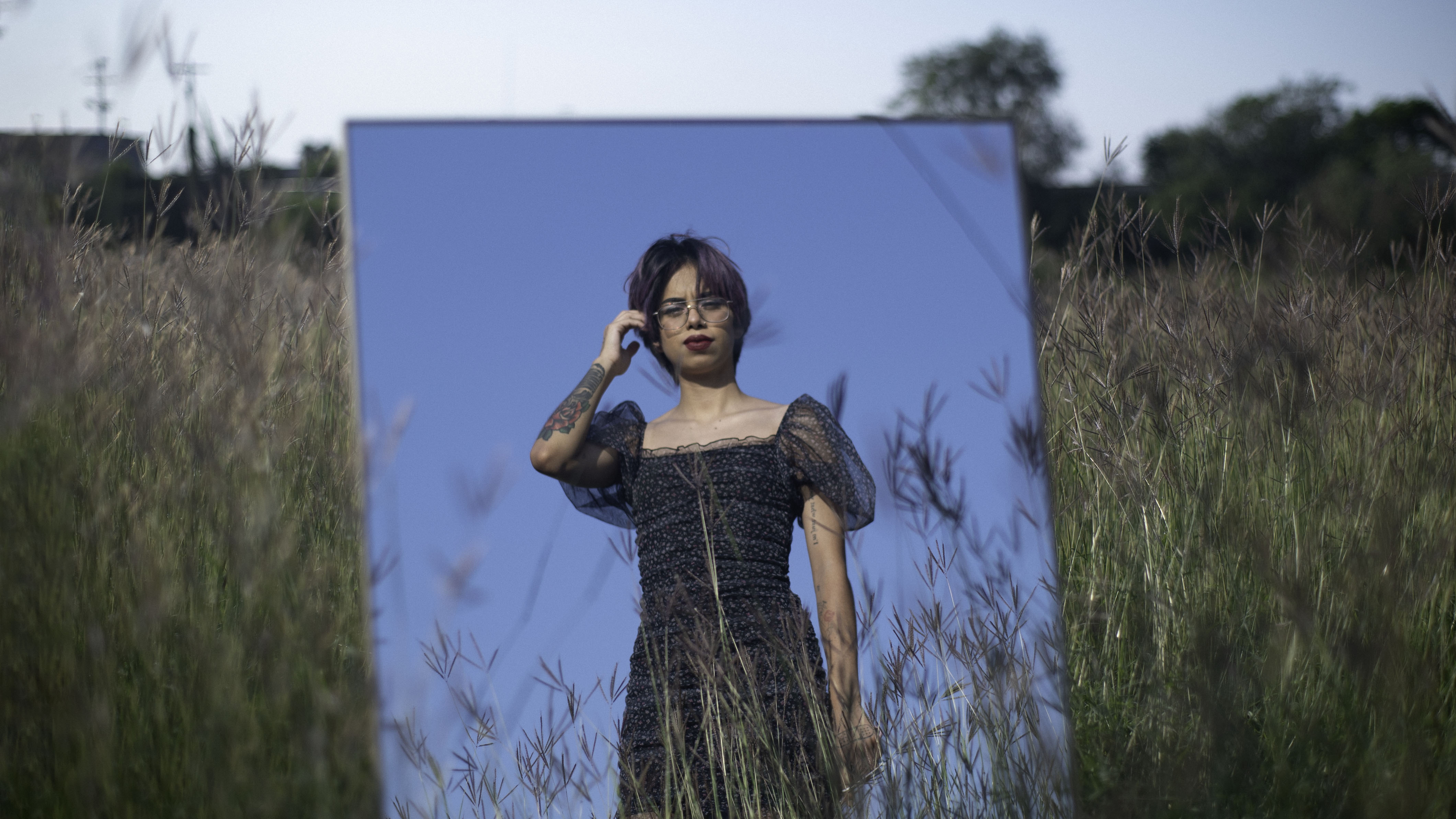 Photo of Estefanía Contreras’s reflection on a mirror. She is wearing a black dress in a field. Her hand is pushing her hair behind her ear. 