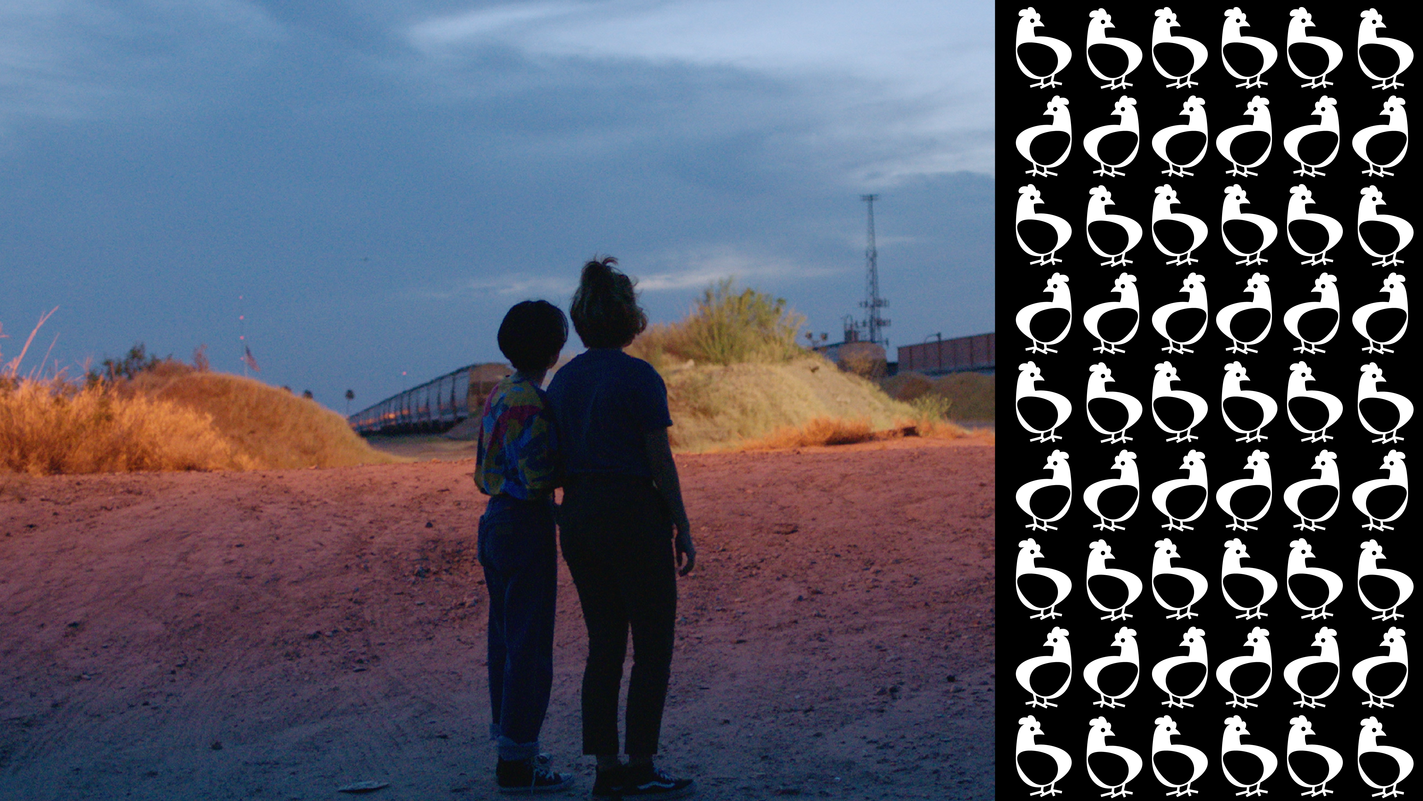 A color still from the film Hummingbirds, in which two people are standing in the foreground on a sandy outdoor landscape with a train approaching from their left. 