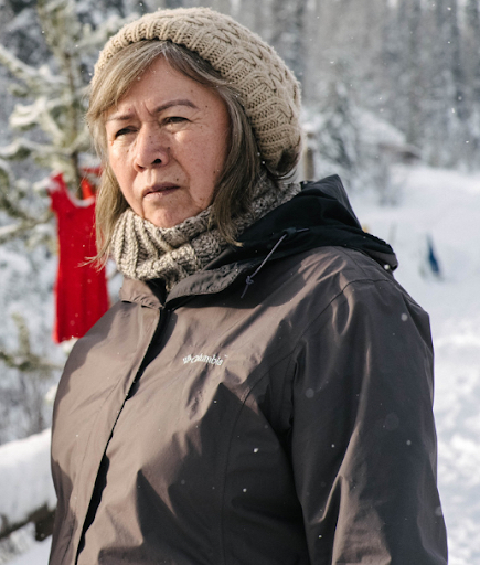 A women in a winter coat and hat looks off as a red dress hangs in the snowy background.