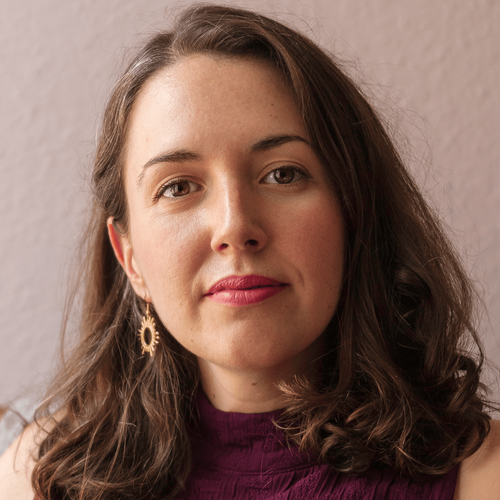 documentary filmmaker Kristine Stolakis looks directly at the camera. her hair is medium brown and falls, slightly curly to her shoulders. A gold sun shaped earring is visible in her right ear. She wears a slight smile. 