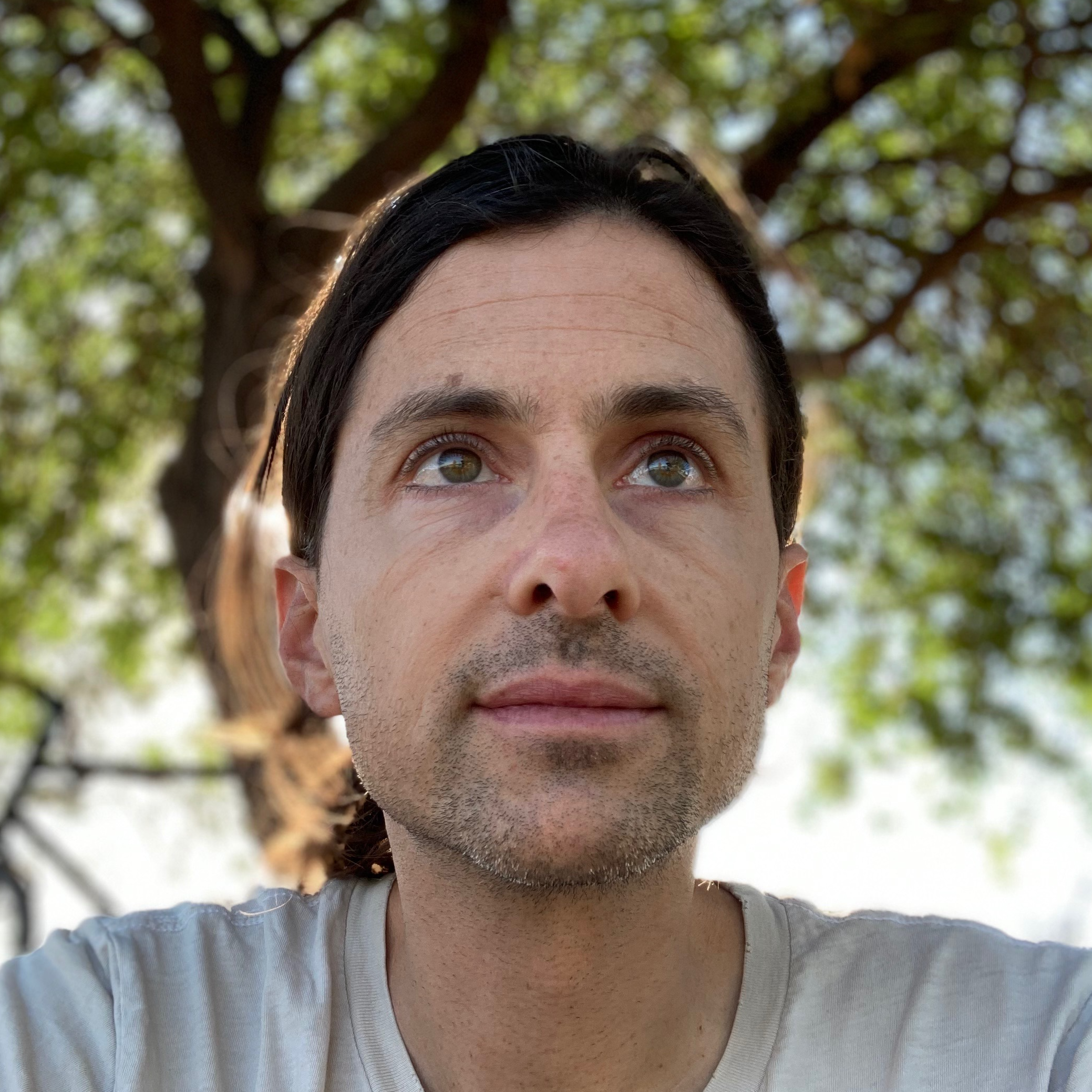 Portrait of Nick Kelso. He wears a shirt and looks away from the camera, in the background there is a tree. The portrait is taken in a low angle. 