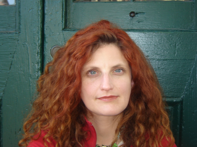 Madeleine Molyneaux, a ginger haired woman with blue eyes, looks directly at the camera. She stands next to a dark green door.