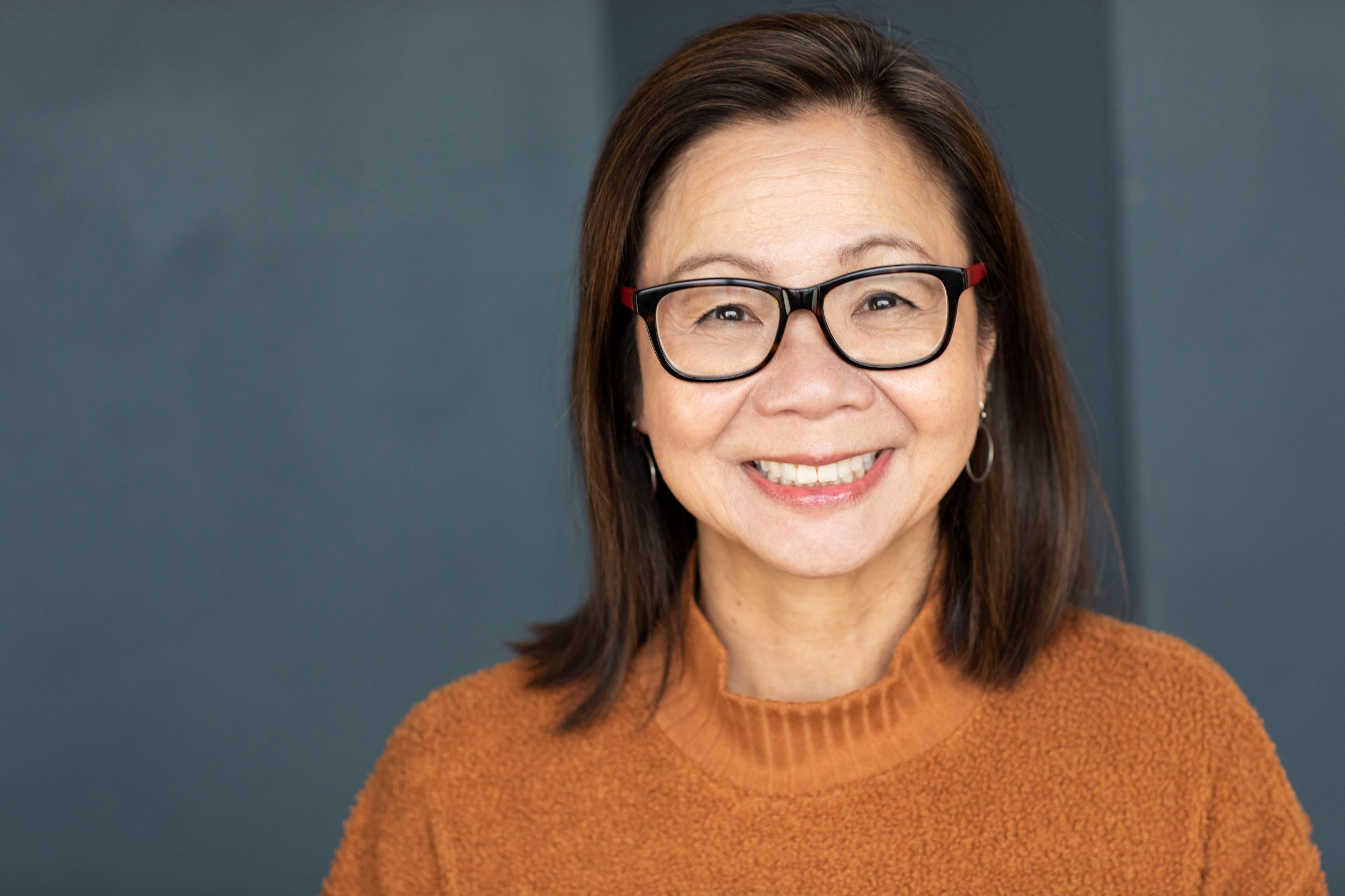 Diane Quon looking directly at camera and is smiling. She has mid-length hair and is wearing a turtleneck, gold hoop earrings and black rimmed glasses. 