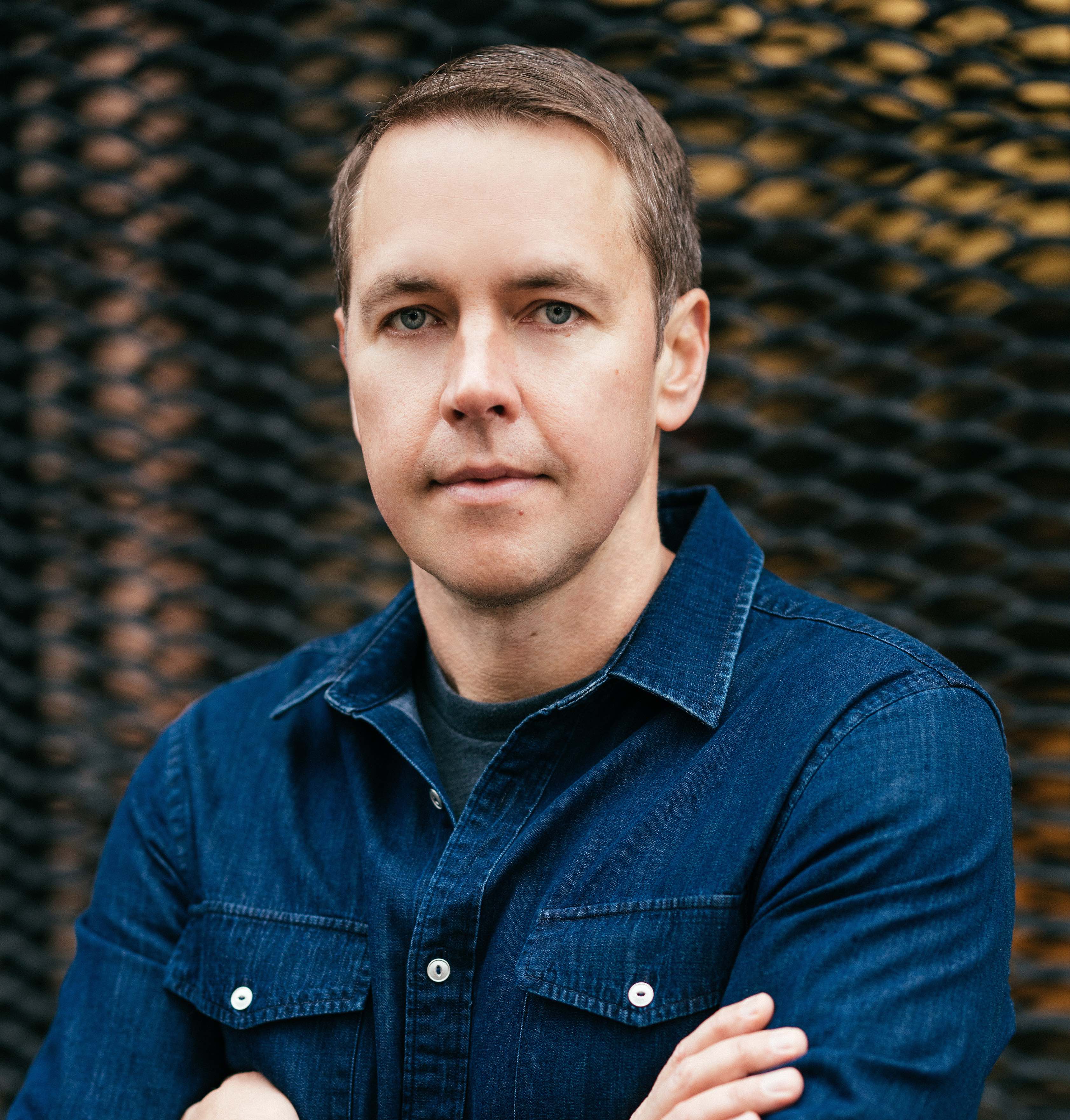 Headshot of a man folding his arms on his chest, he wears a long sleeve shirt and short hair, he looks at the camera