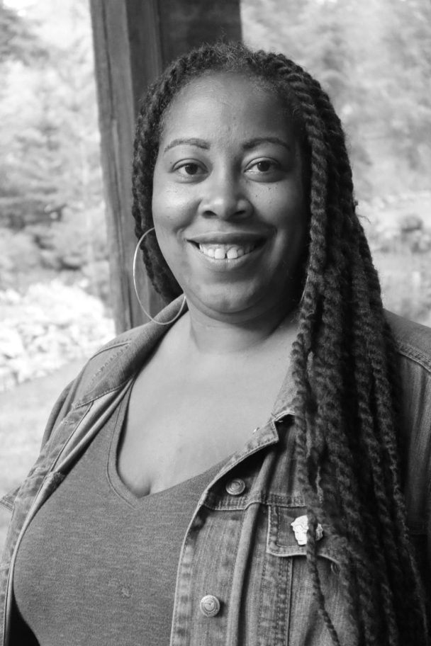 Angela Tucker looking directly at the camera and smiling. She is wearing a denim jacket, a turquoise shirt, and purple and brown crocheted dreads. Black and white portrait