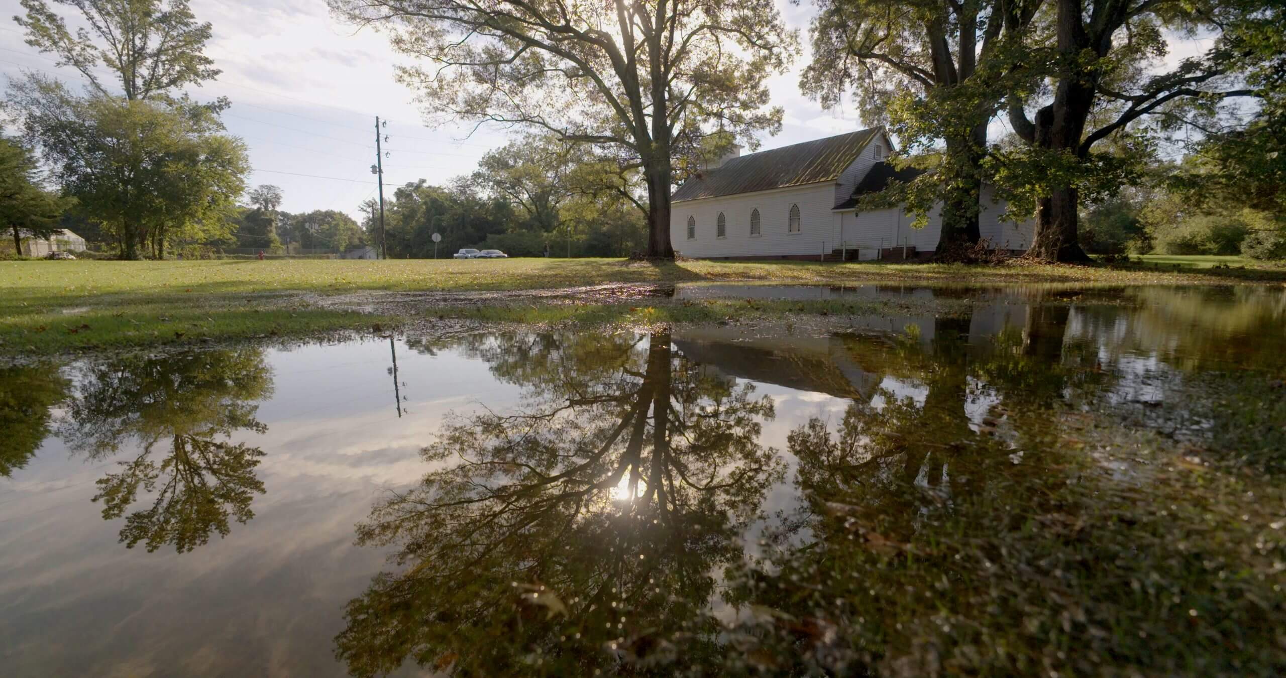 A large tree is shown reflected in a large puddle with a white church in the background.