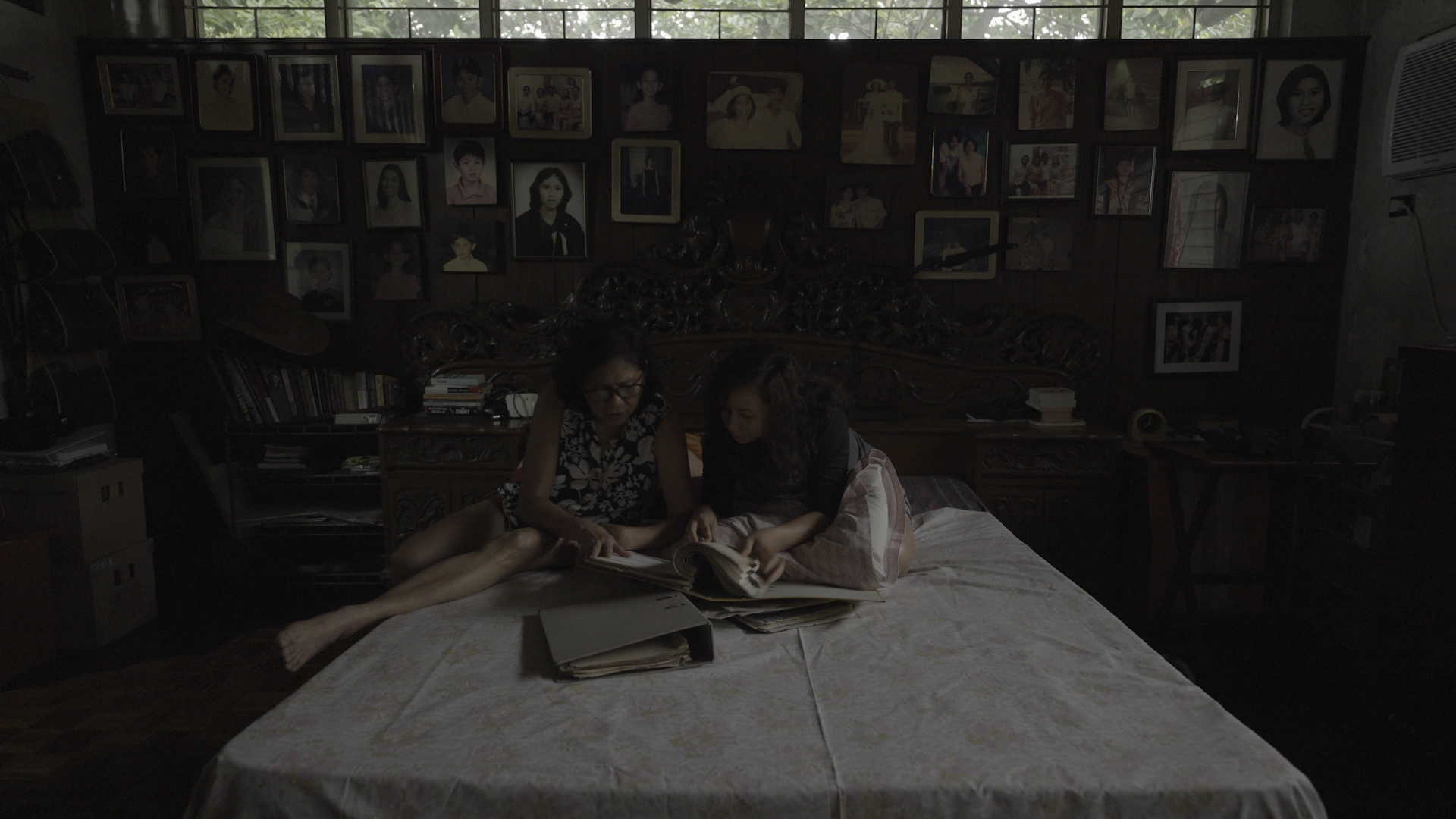 Two people sit on a bed pouring over photo albums and books in a dimly lit room. The wall behind them is filled with family portraits.