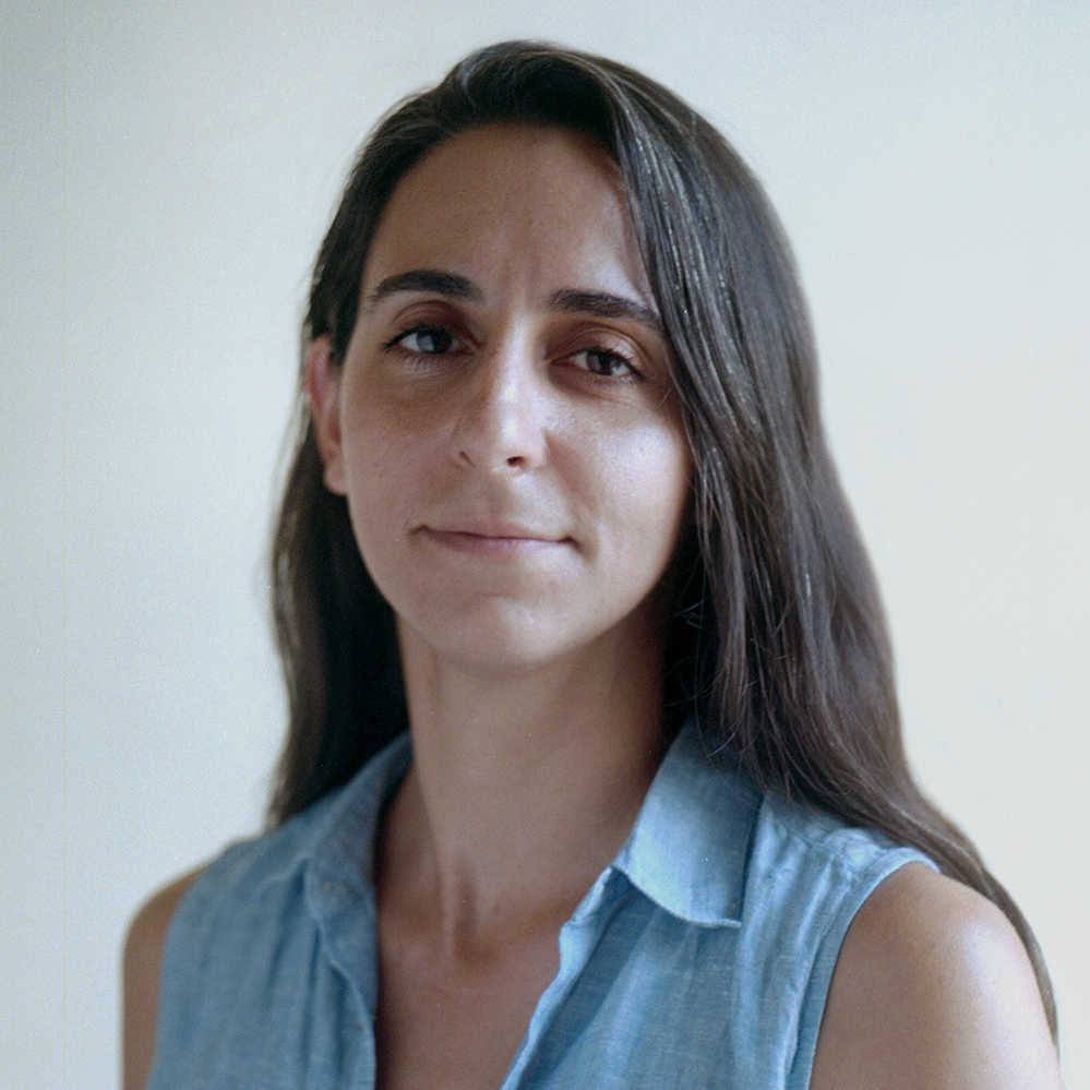 A photograph of a young woman with long dark brown hair smiling. She is wearing a blue sleeveless collared shirt, and sitting in front of an off-white background. The photograph is cropped at the shoulder.