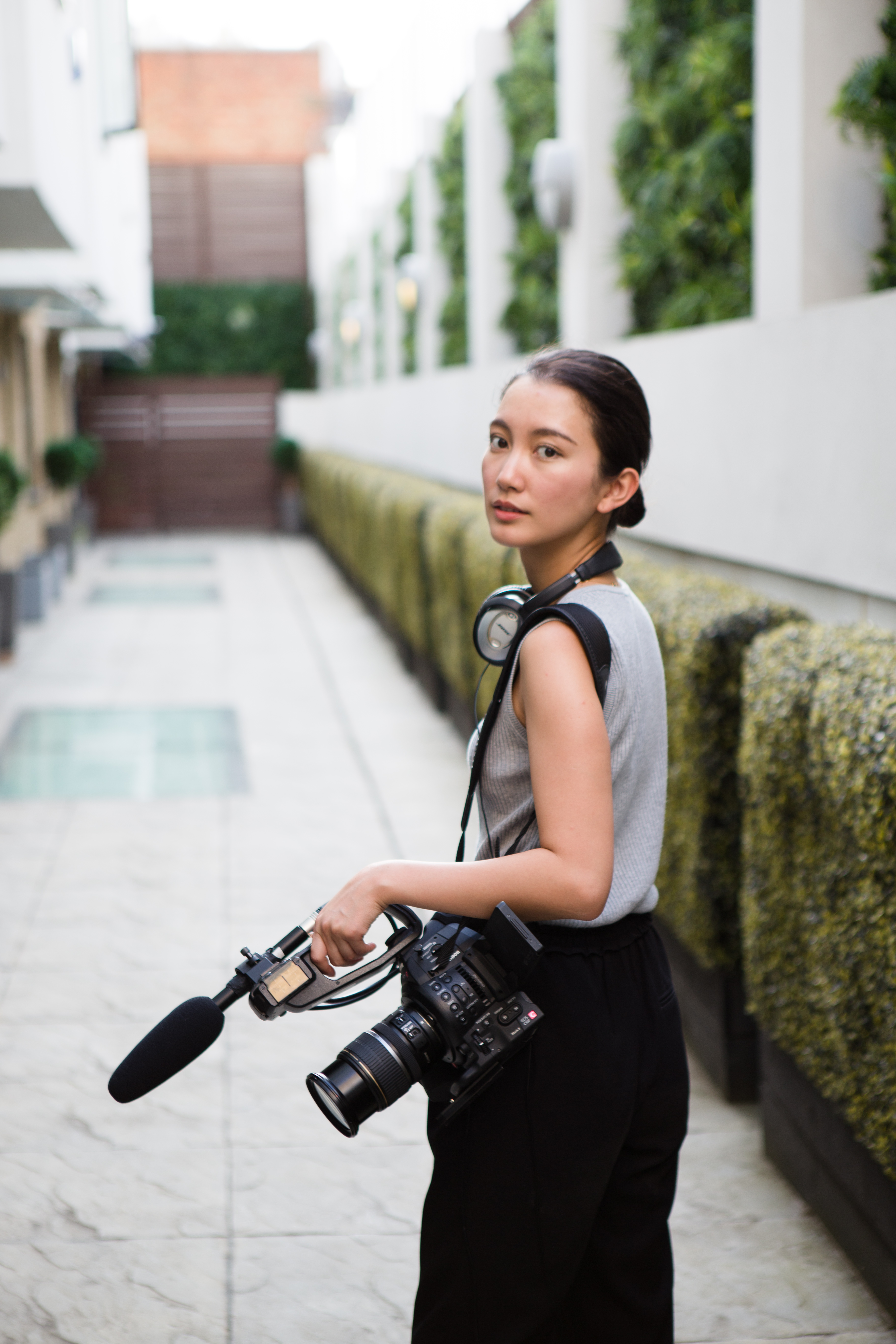 A woman, dark brown hair, standing outside with a film camera over her left shoulder, she wears headphones around her neck. 