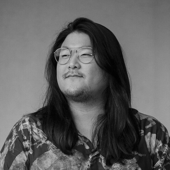 An Asian American man with wearing wire rimmed glasses and shoulder length dark hair, looks off to the left of the photo frame. Headshot of Board of Directors member David Lee.