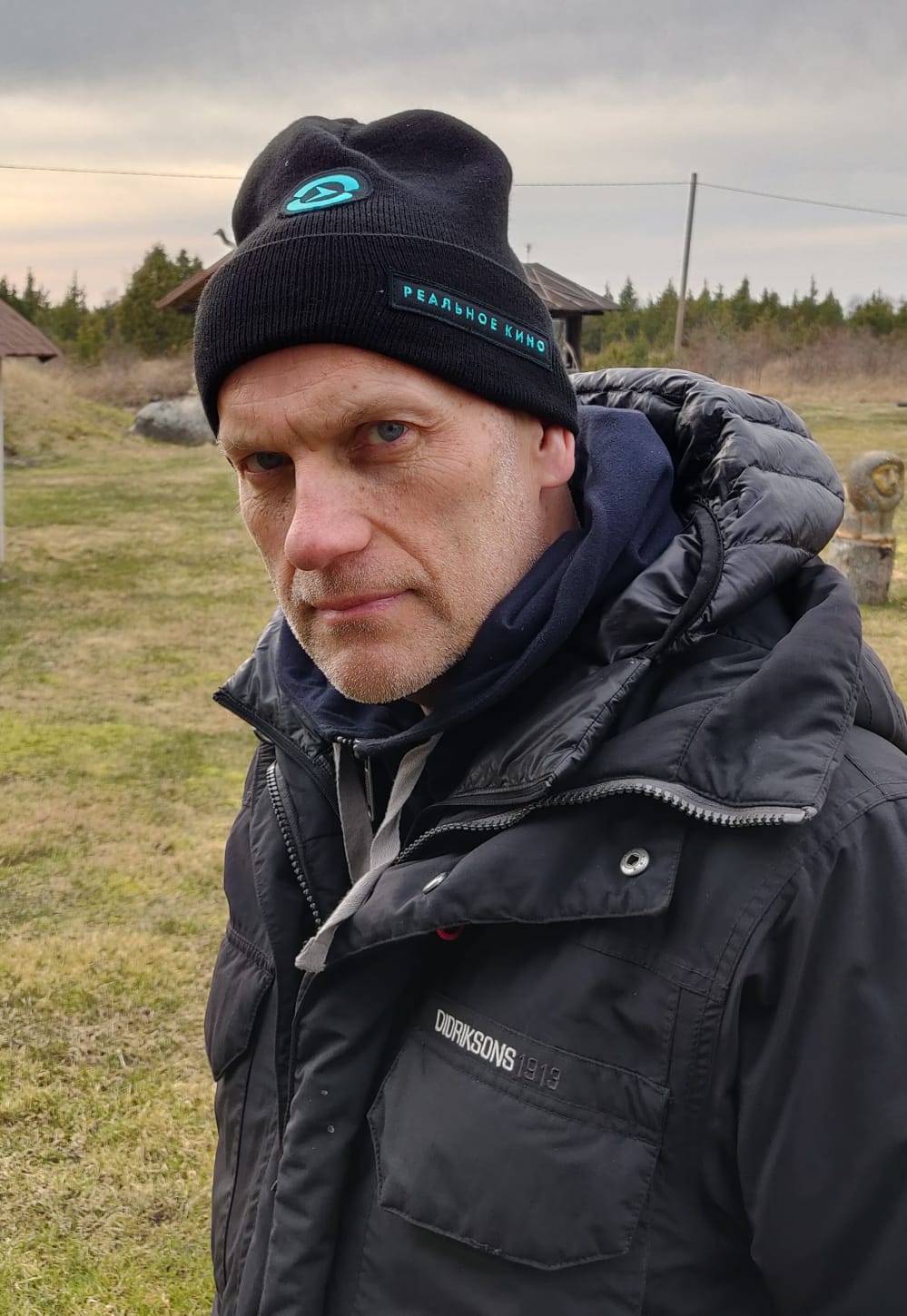 Portrait of a man wearing a hat standing in an open field, he wears a jacket and looks seriously at the camera