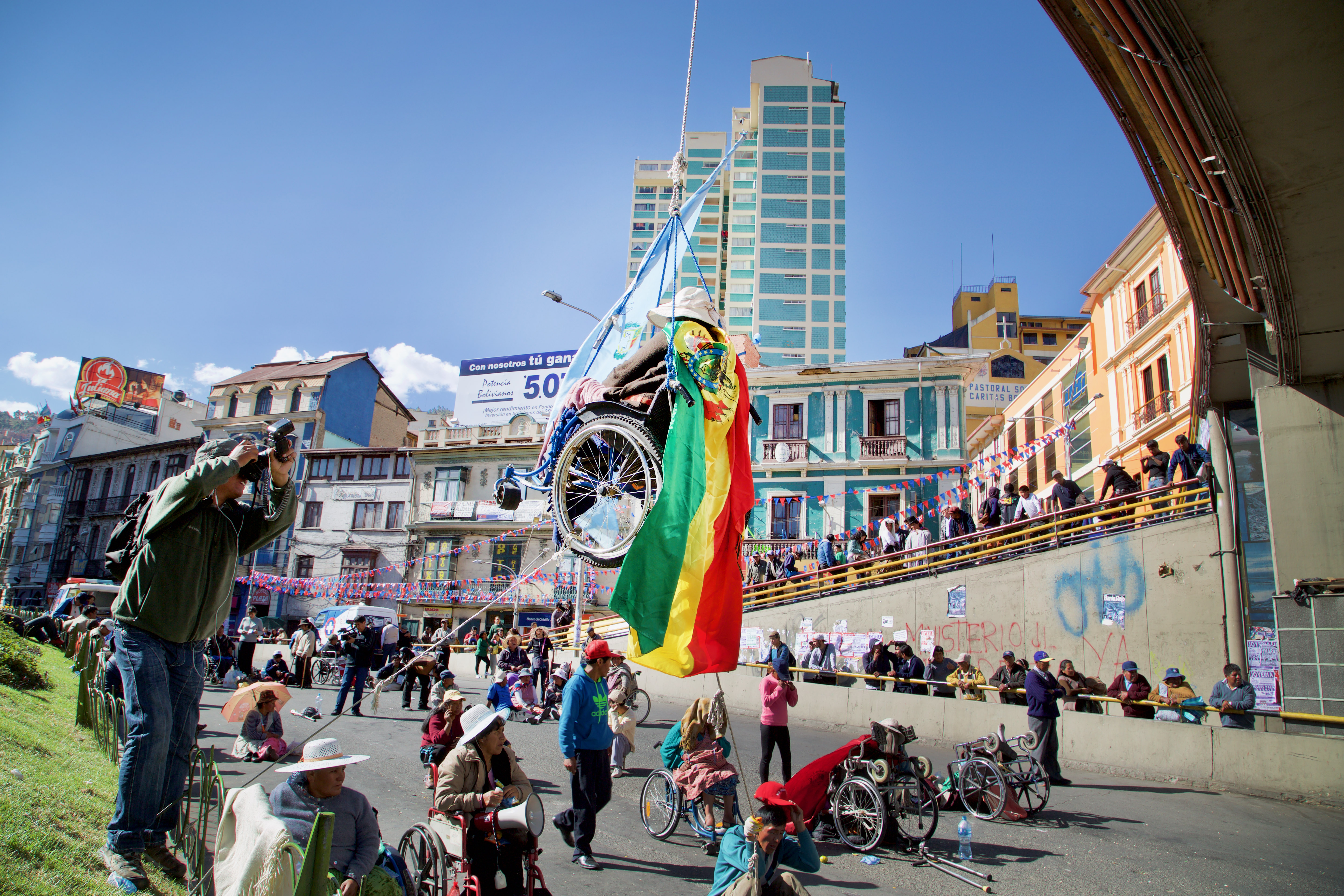 Still from La Lucha. Leader Rose Mery Guarita is suspended in her wheelchair from a bridge over the busiest street in La Paz, with a Bolivian flag draped over her. Below her, fellow protesters have their wheelchairs turned around, facing away from the bridge. The bustling scene captures the intensity of the protest, with buildings and a high-rise tower forming the backdrop. A photographer is positioned on a grassy area to the side, capturing the moment that symbolizes the determination of the movement.