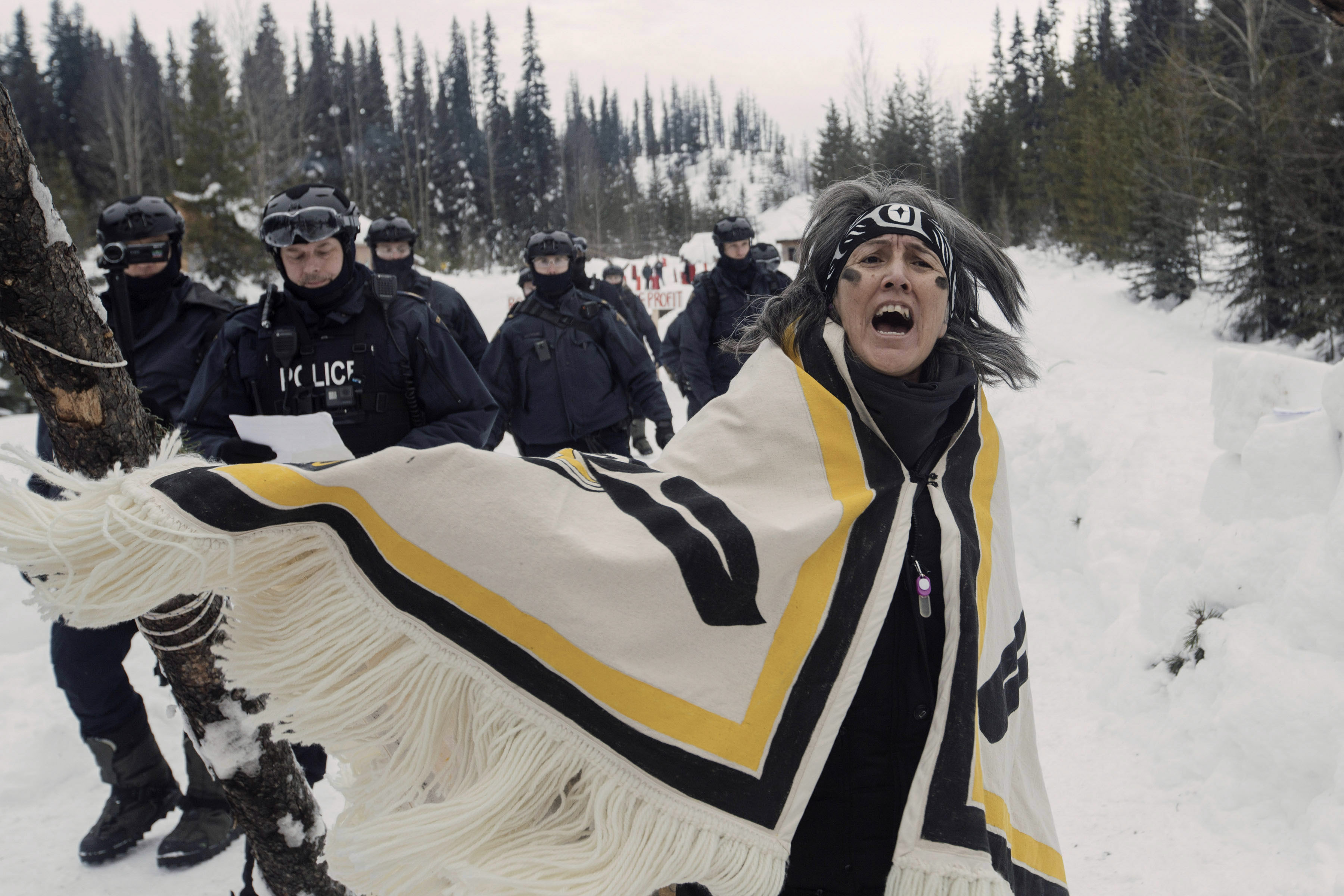 Still from Yintah. Against a snowy, tree-lined landscape, a woman with charcoal on her face sings and dances while police approach from behind.