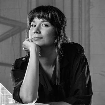 Lisa Jackson looking directly at the camera. She is leaning her chin on her hand. The background is out of focus and has shadows on the wall. Black and white portrait