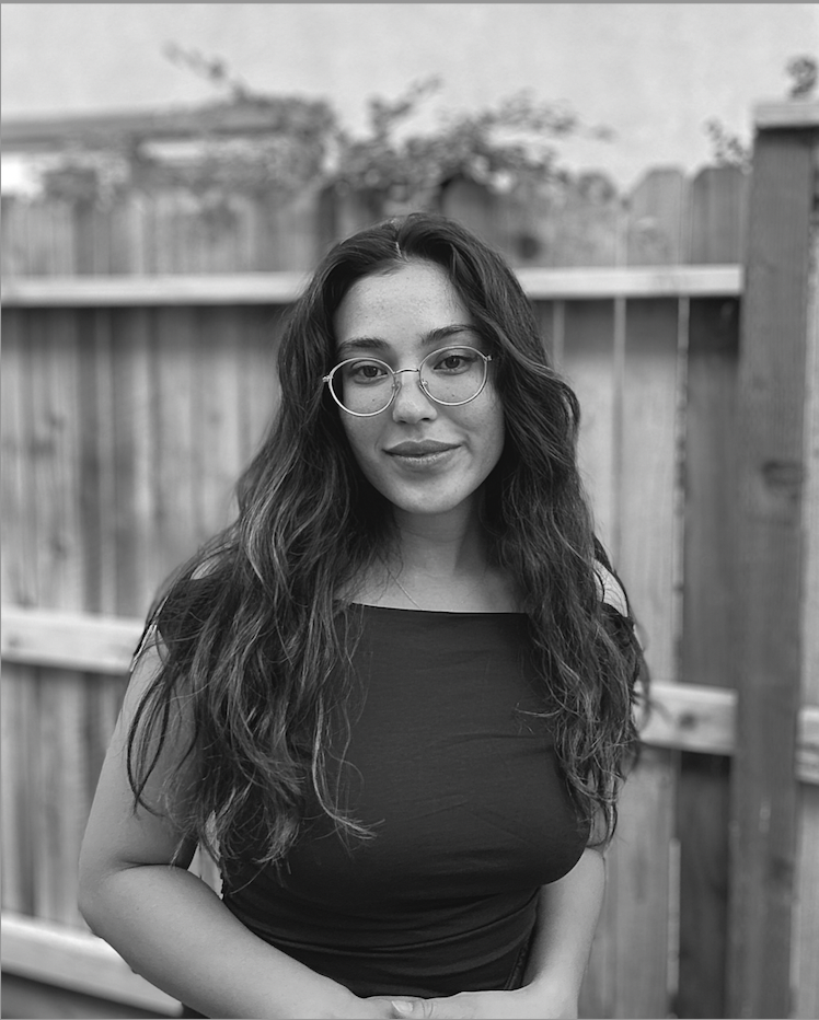 A mixed race woman stands in front of a wooden fence. She has long, dark hair and wears wire rimmed glasses and smiles slightly while looking directly at the camera. A headshot for filmmaker Bree Nieves Robert. 