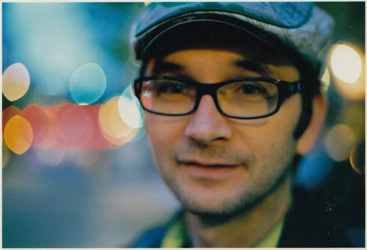 David Redmon looking straight ahead. He wears dark-rimmed glasses and a flat cap. Black and white portrait, with out of focus background.