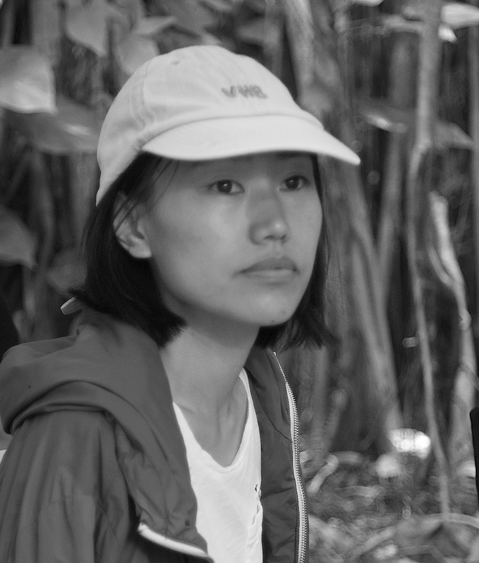 Portrait of Sarah Kim, she looks away at the camera, wears a shirt and a hoodie, and a cap. Her hair is short. In the background there are plants.