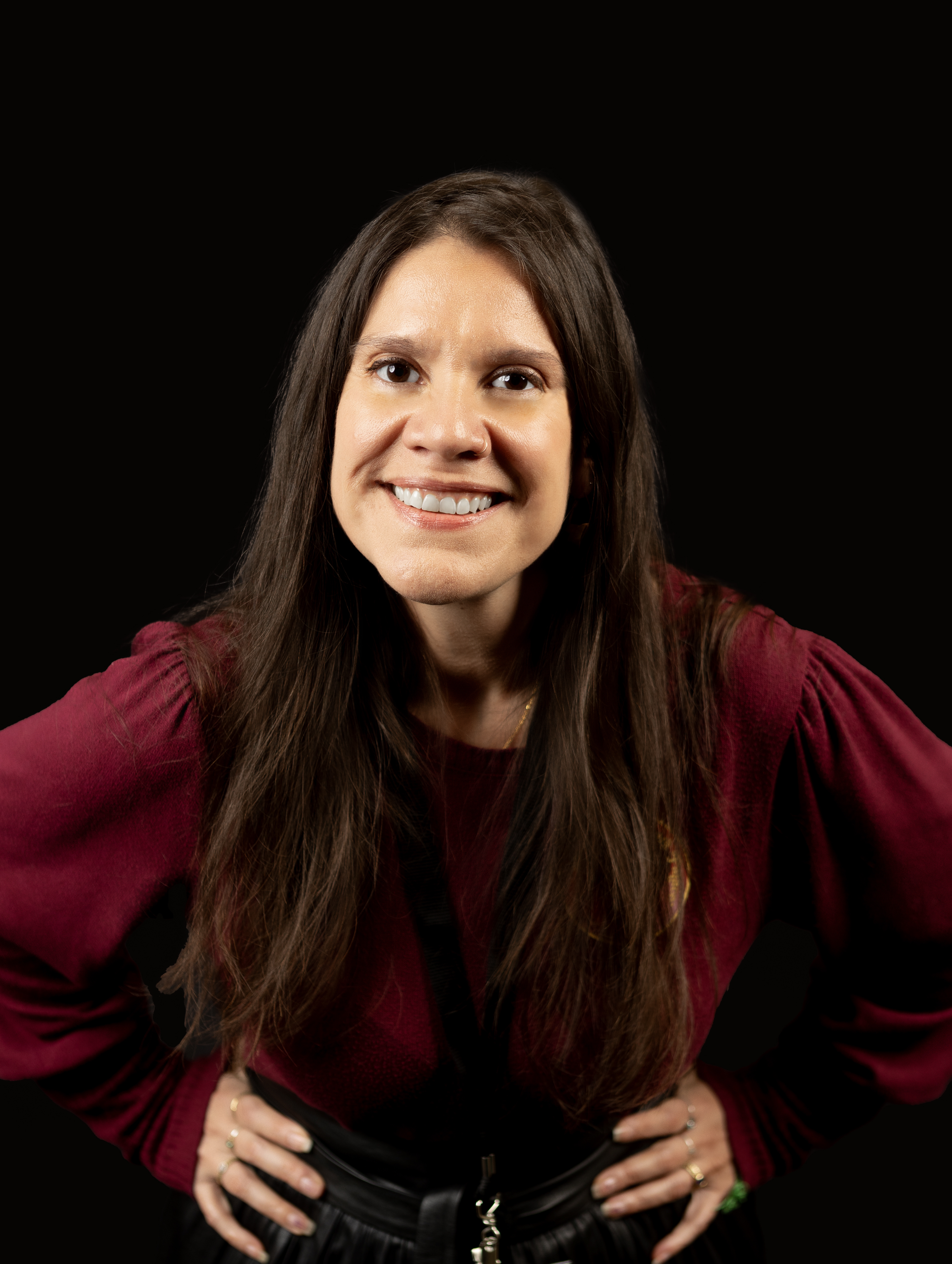 Hazel is a Colombian woman with long brown hair and brown eyes and a bright smile, leaning forward, hands on hips, and wearing a long-sleeved maroon sweater.