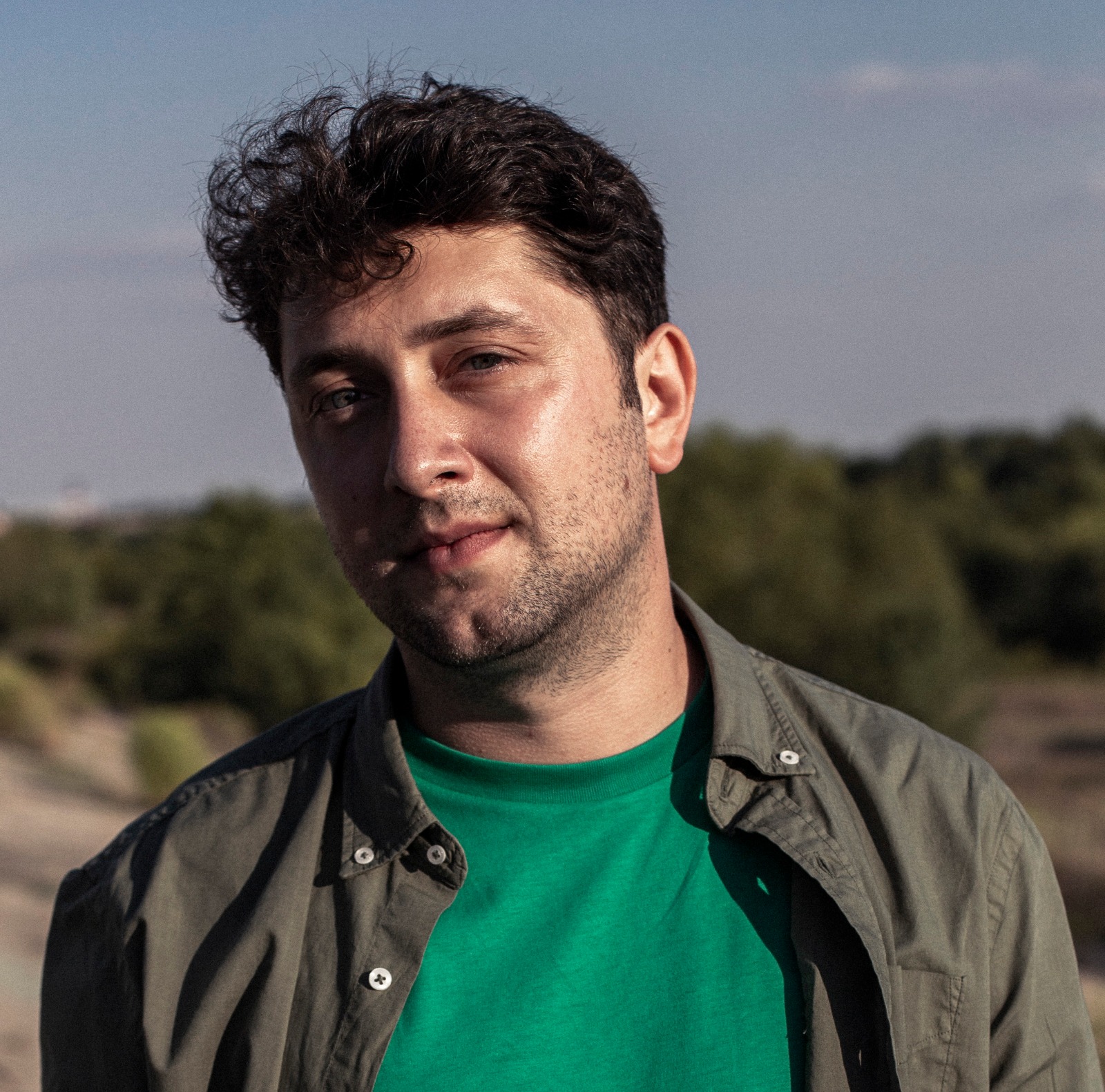 Portrait of a man with short hair and his head tilted to the right, he wears t-shirt under a shirt, the background is an open air forest landscape