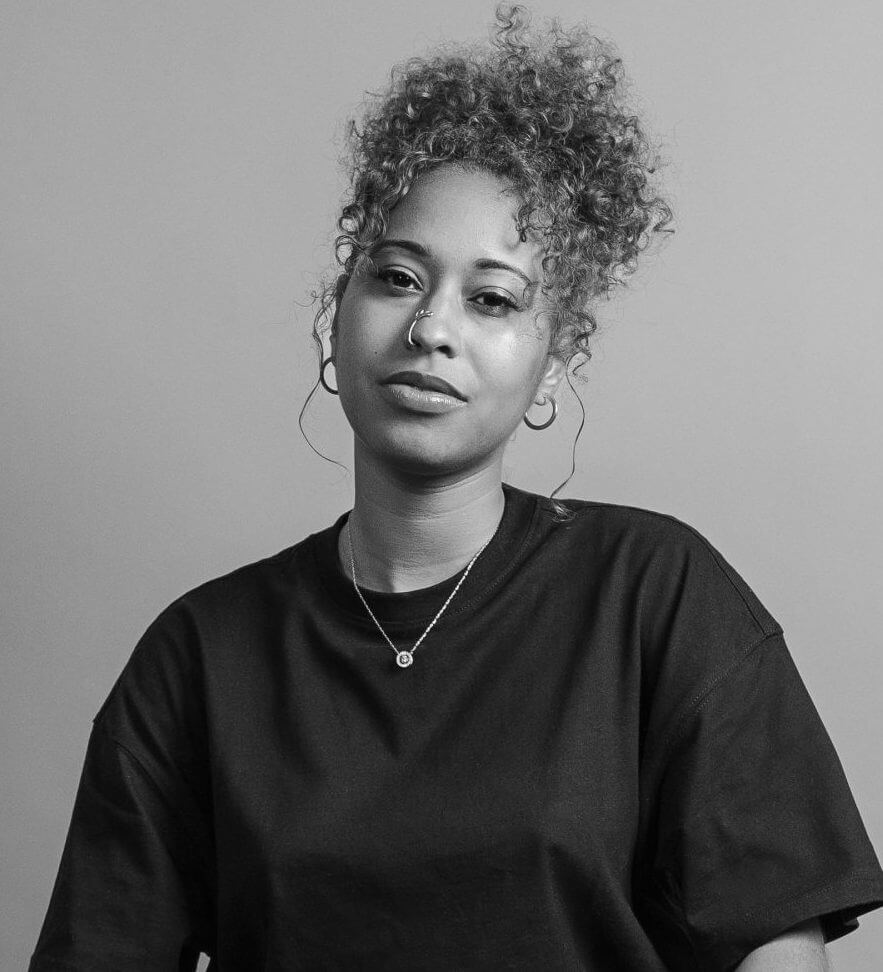 Black woman with curly, brown hair sits in front of a tan backdrop on a stool with legs crossed, wearing a black shirt, Black loafers and white socks. Black and white portrait.