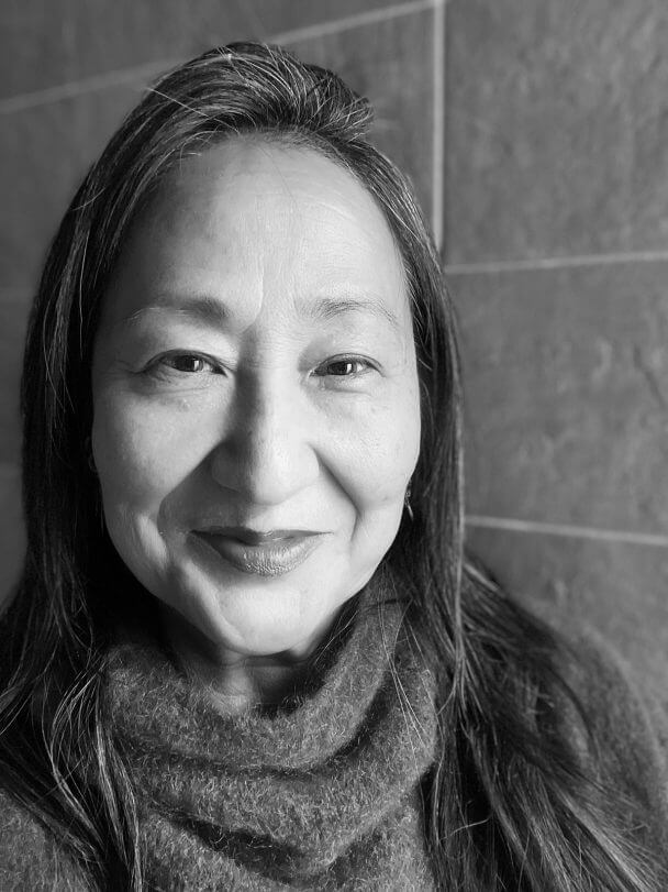 Rea Tajiri looking directly at the camera. She wears a red sweater with red lipstick and is smiling. Black and white portrait