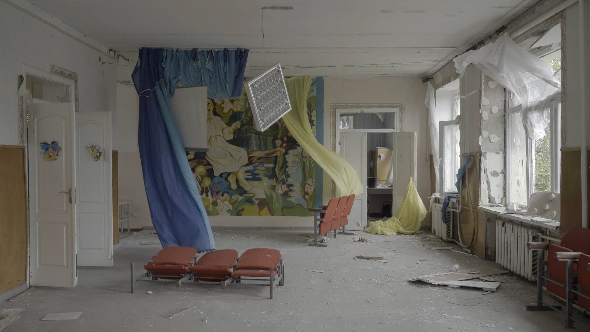 Production still of Intercepted. A ransacked classroom in a school near Mikholaiv is shown. Blue and yellow cloths are hanging from the ceiling, a light fixture is hanging from the ceiling by a thread. The classroom door is wide open and wind is blowing from the destroyed windows