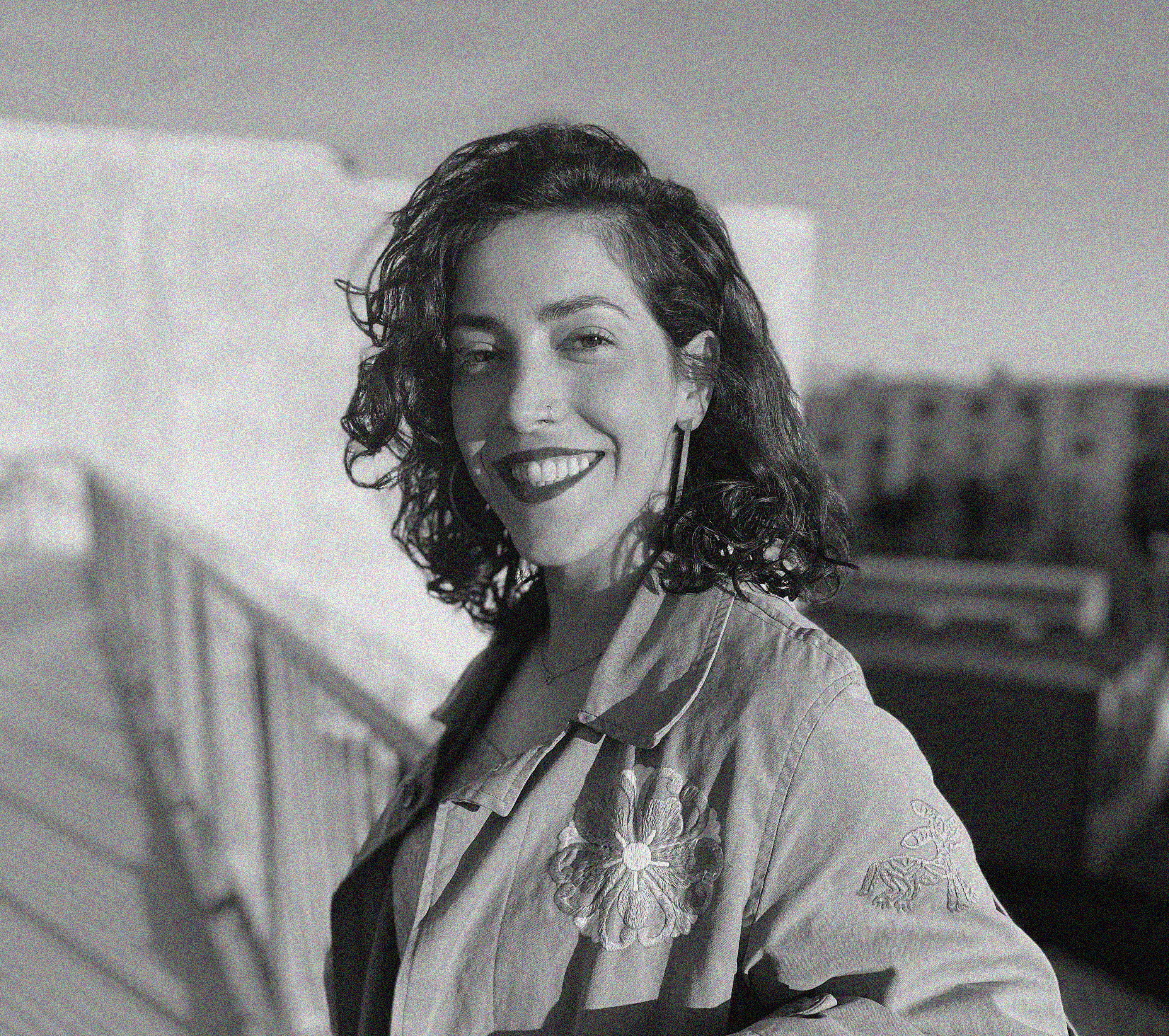 A black-and-white photograph of an Arab woman with curly hair and a smile, standing outdoors on a sunny day. She is wearing a light jacket with floral embroidery and large hoop earrings. The background features a blurred urban landscape.