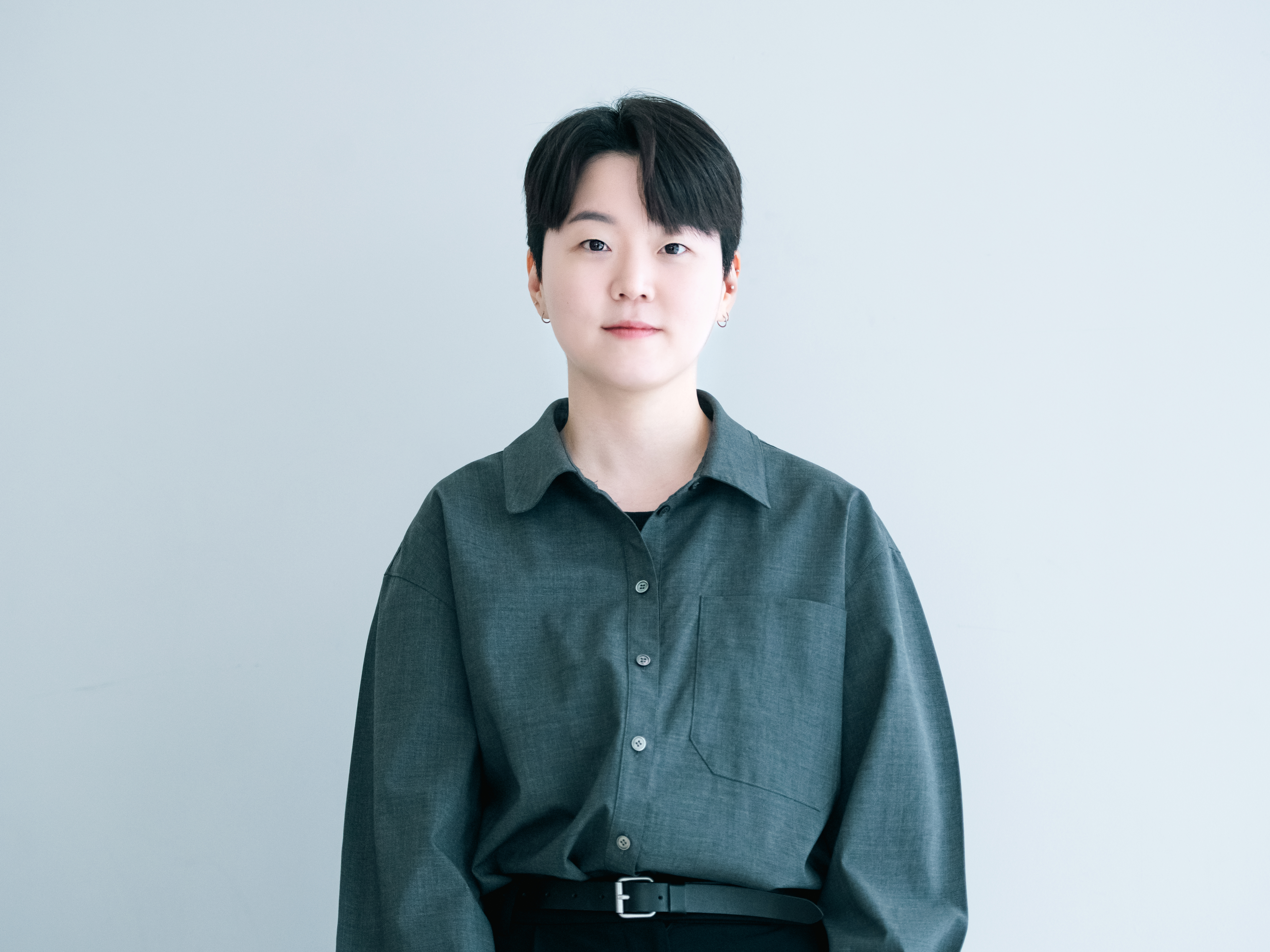 The director Minji Ma is facing forward with a slight smile against a white background. They have black hair and a round Asian face with a short haircut. They also have very light skin tones. They have multiple piercings on their ears. They are wearing a gray shirt tucked into black slacks, accessorized with a belt.