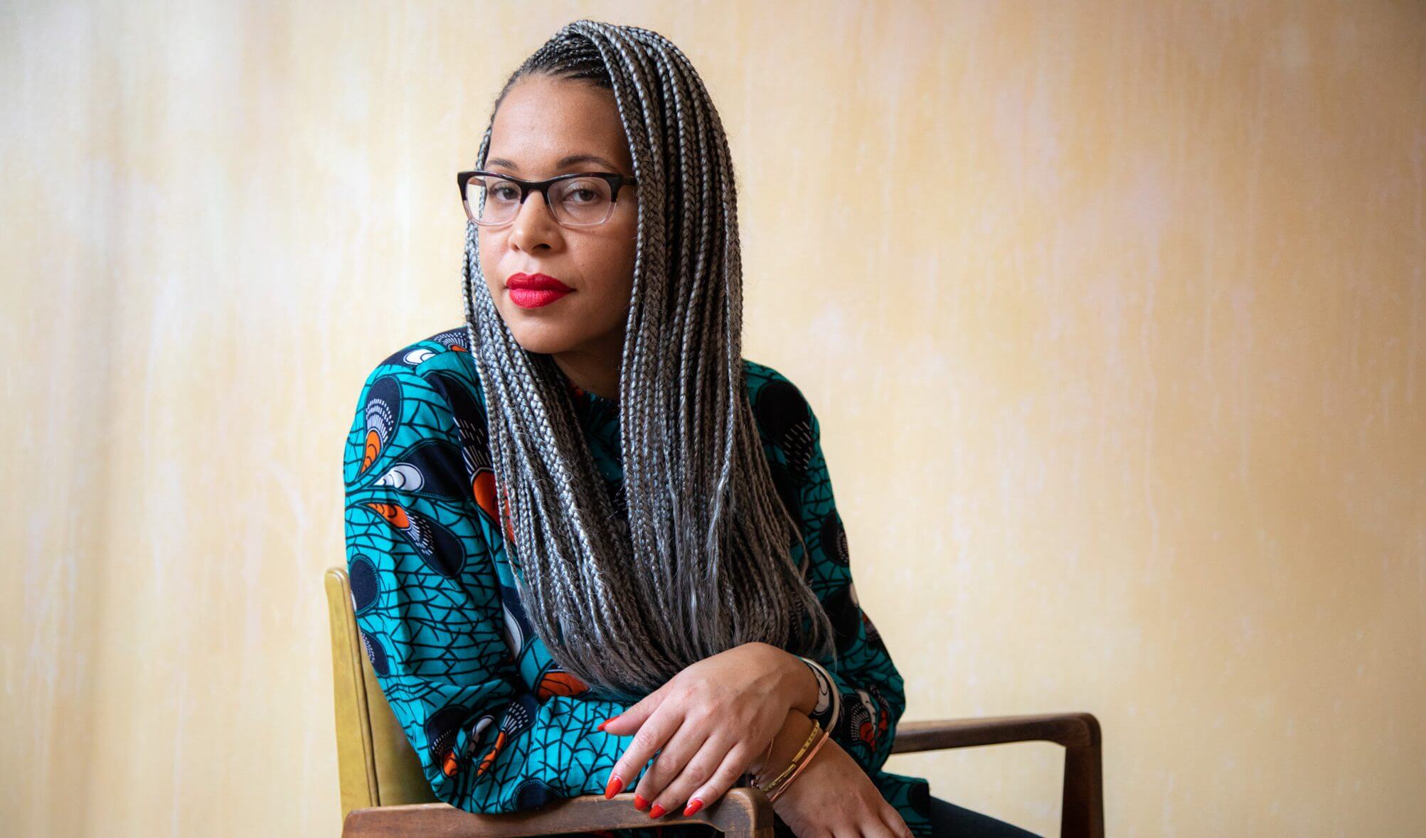 Loira Limbal is sitting in a chair, she looks at the camera with her head slightly turned. Loira wears lipstick and her hair is braided. Portrait in black and white.