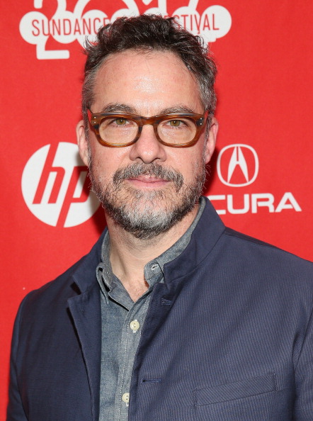 Portrait of Tony Gerber. Tony is in front of a photo call, he wears a jacket, a buttoned shirt, and thick frames. He has short heard, beard, and mustache. He looks directly at the camera and smiles slightly.