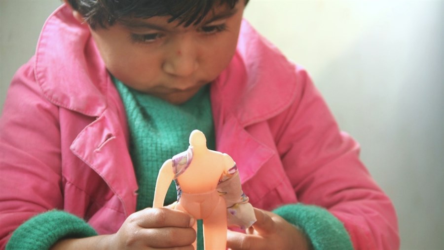 A young child wearing a pink jacket holds a doll with a missing head and wrapped in bandages close to their face and investigates it.