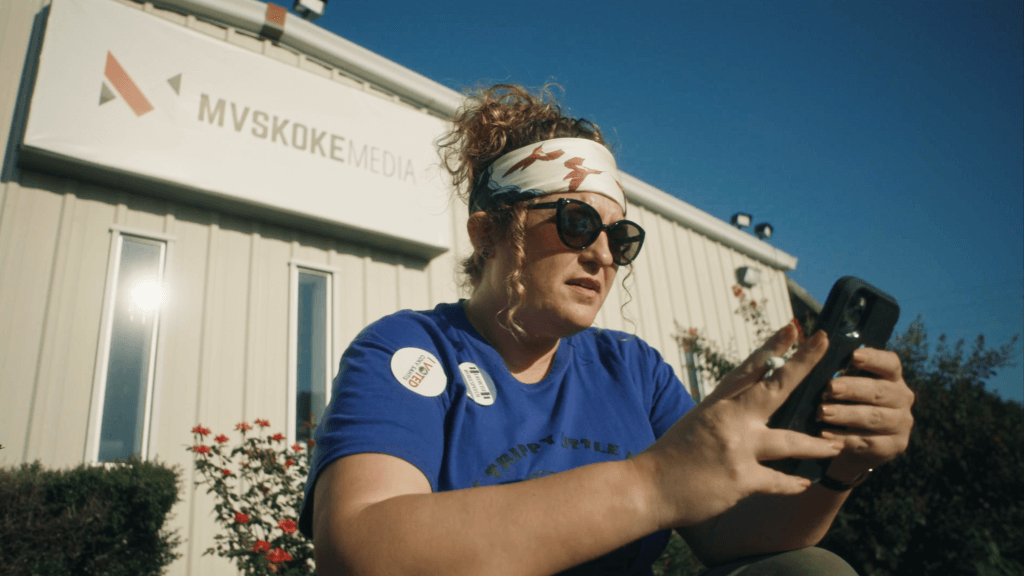 Indigenous journalist Angel Ellis sits in front of Mvskoke Media wearing a bandana , sunglasses and a "I Voted" sticker on her blue T-shirt. She's smoking a cigarette while scrolling through her cell phone on a warm summer evening.