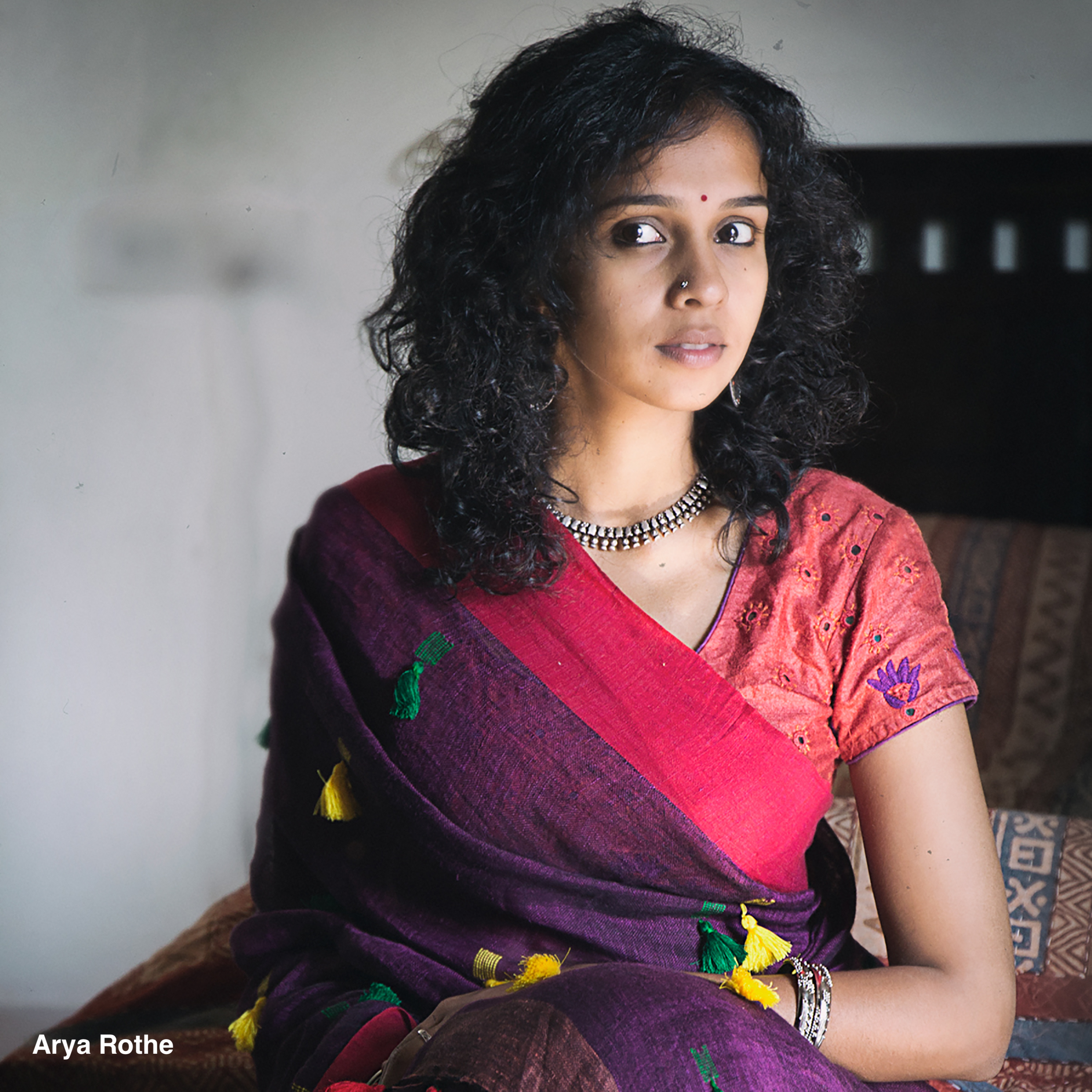 Headshot of a curly black-haired woman dressed in a colorful sari against a white wall.