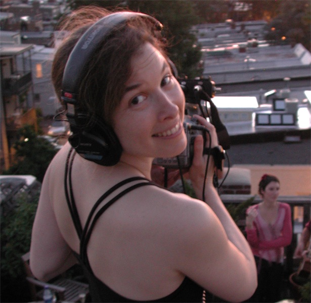 A portrait of a smiling woman wearing headphones and holding a camera.