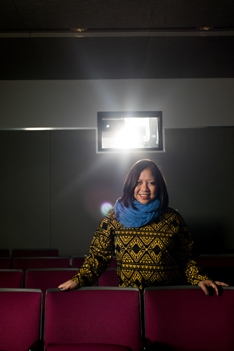 Ramona Díaz standing in an empty movie theatre, wearing a yellow and black sweater with a blue scarf. She has dark shoulder length hair, and is smiling.