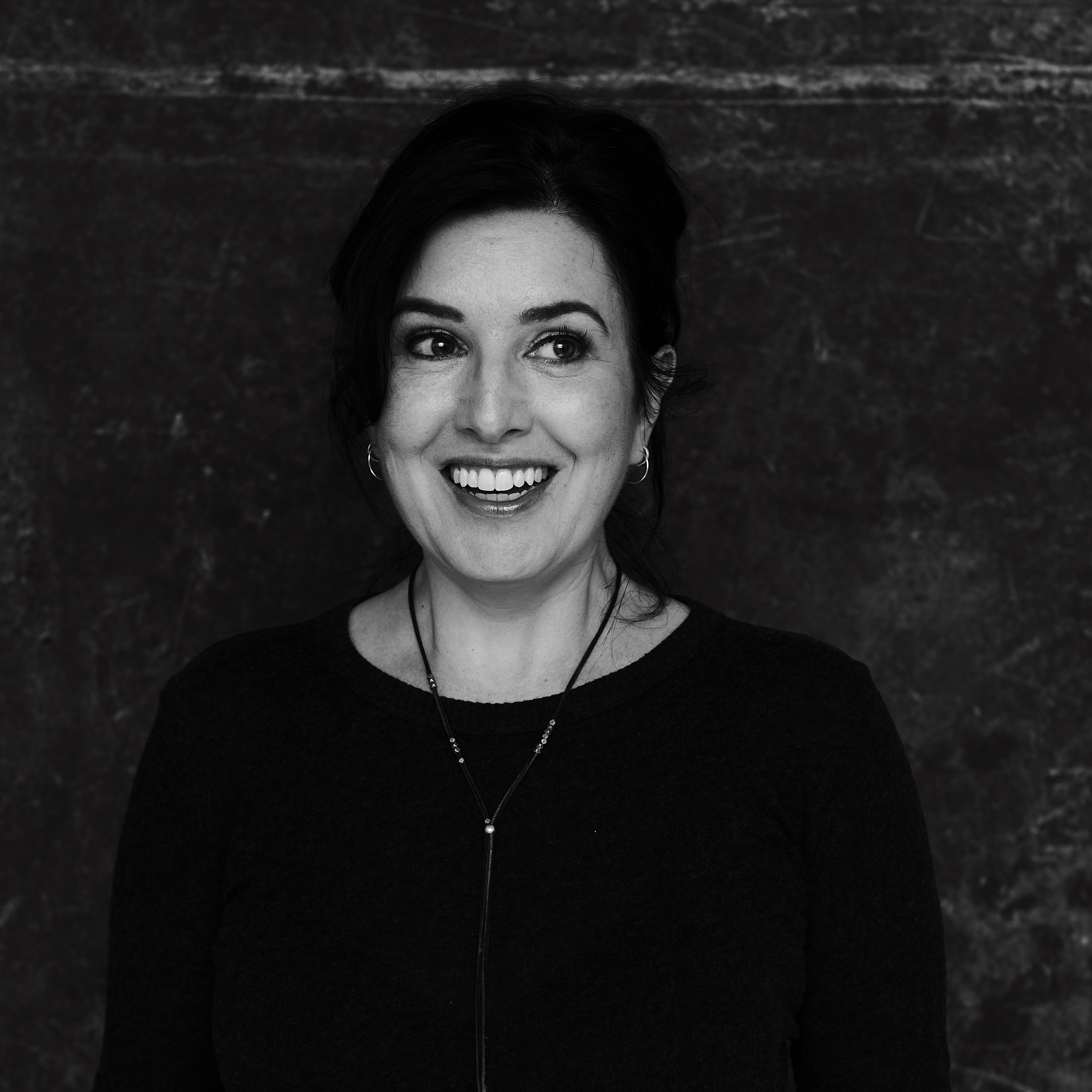 A black and white photo from the torso up of a woman wearing a black sweater with a lanyard necklace, and dark hair pulled back, smiling.