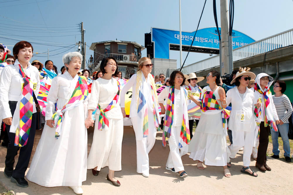 A group of many women walking dressed in white with a colorful scarf. They walk past a watch tower and a wired fence.