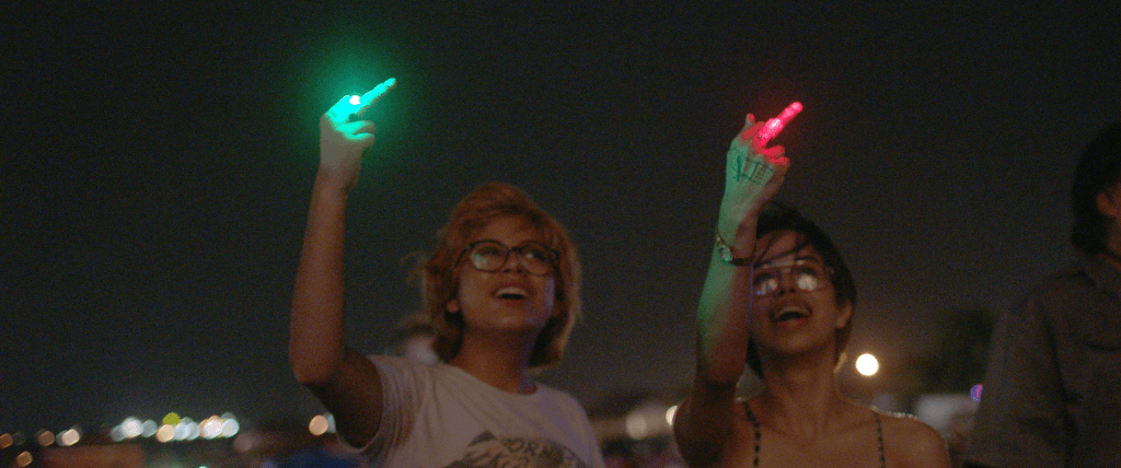 Silvia and Beba smile as they point their middle finger at the firework-lit sky. There is a red light on Beba's middle finger, and a green light on Silvia's.