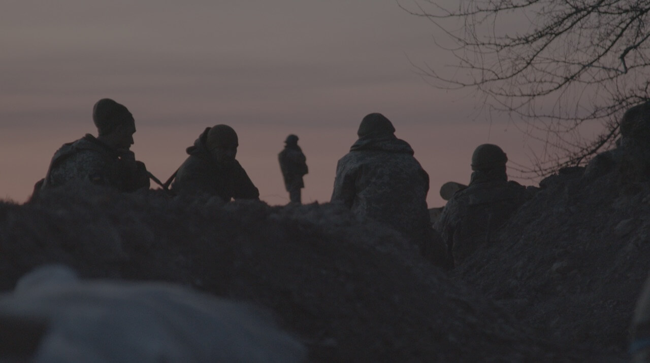 Still from Frontline. We see soldiers sitting on the ground on the edge of a trench. We see only their backs as they are looking towards pink, evening sky. Only one of the soldiers is standing up, observing the horizon.