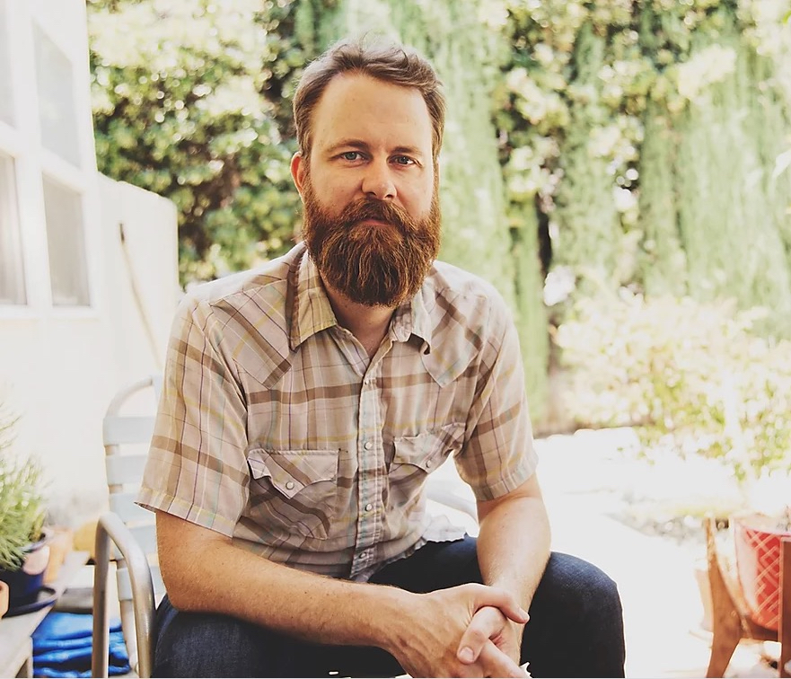 A bearded white man looks directly at the camera, arms crossed, and smiles.